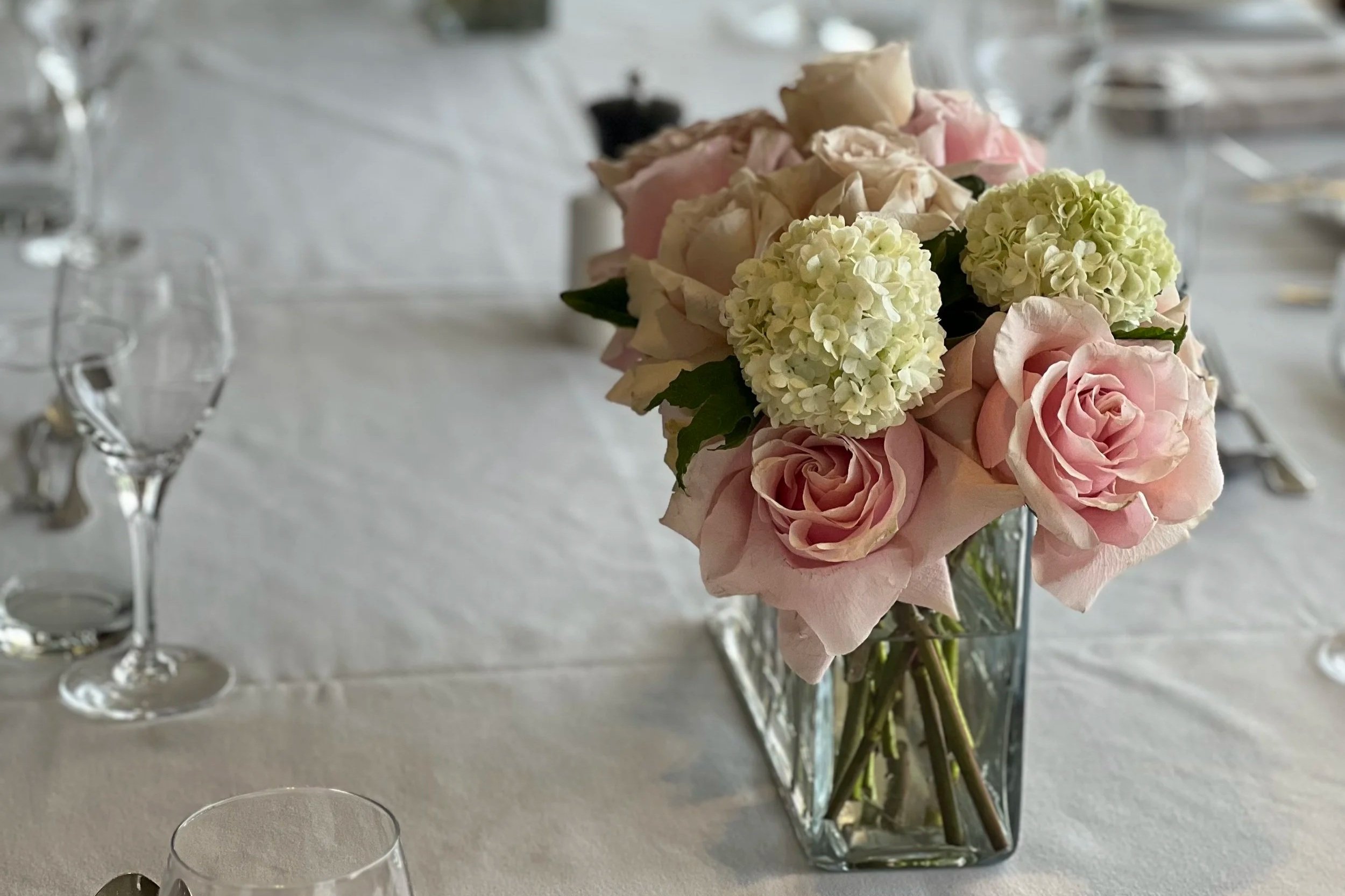 A floral centerpiece with pink roses, white hydrangeas, and peach-colored flowers in a glass vase on a table.
