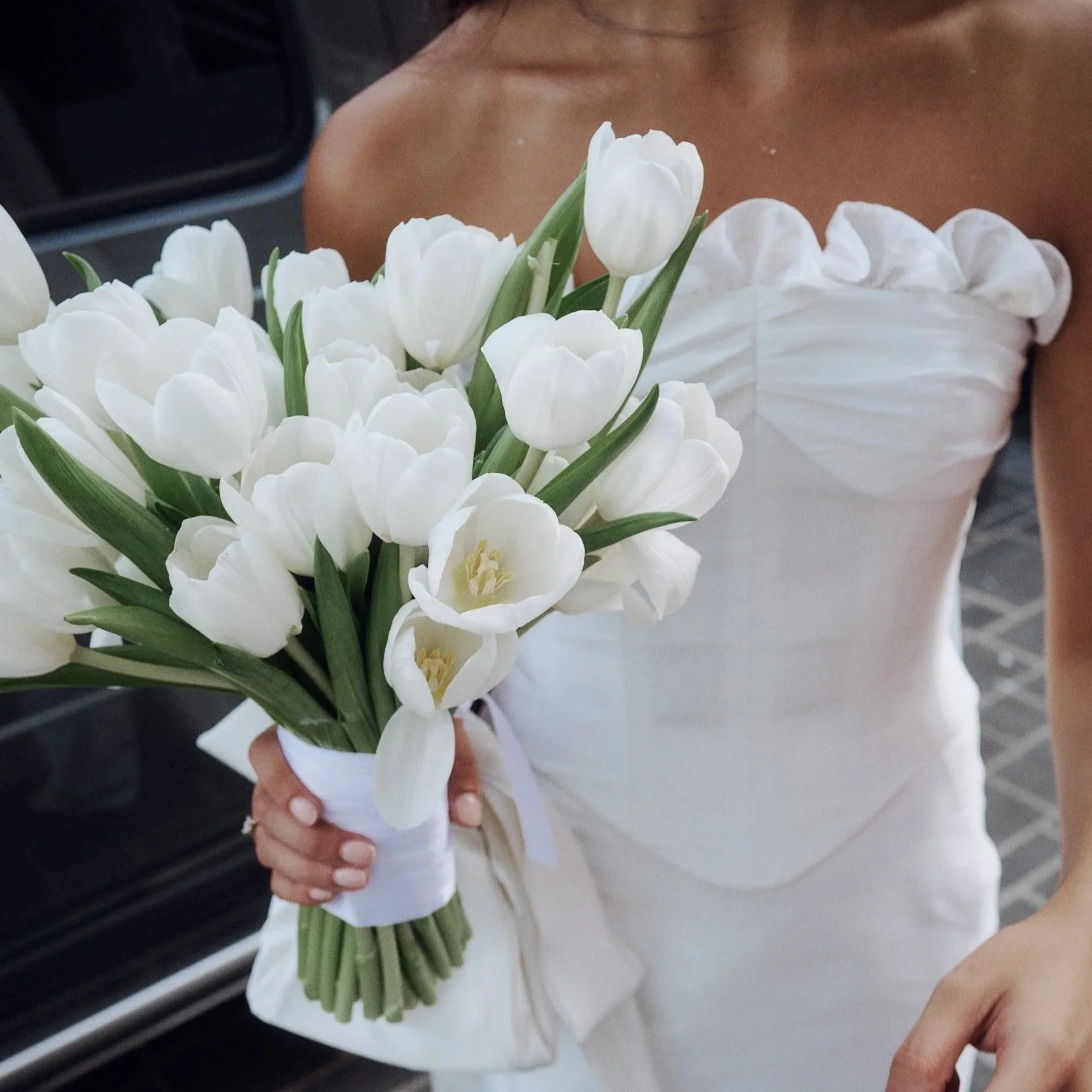 A woman in a white strapless dress holding a bouquet of white tulips with green stems and leaves.