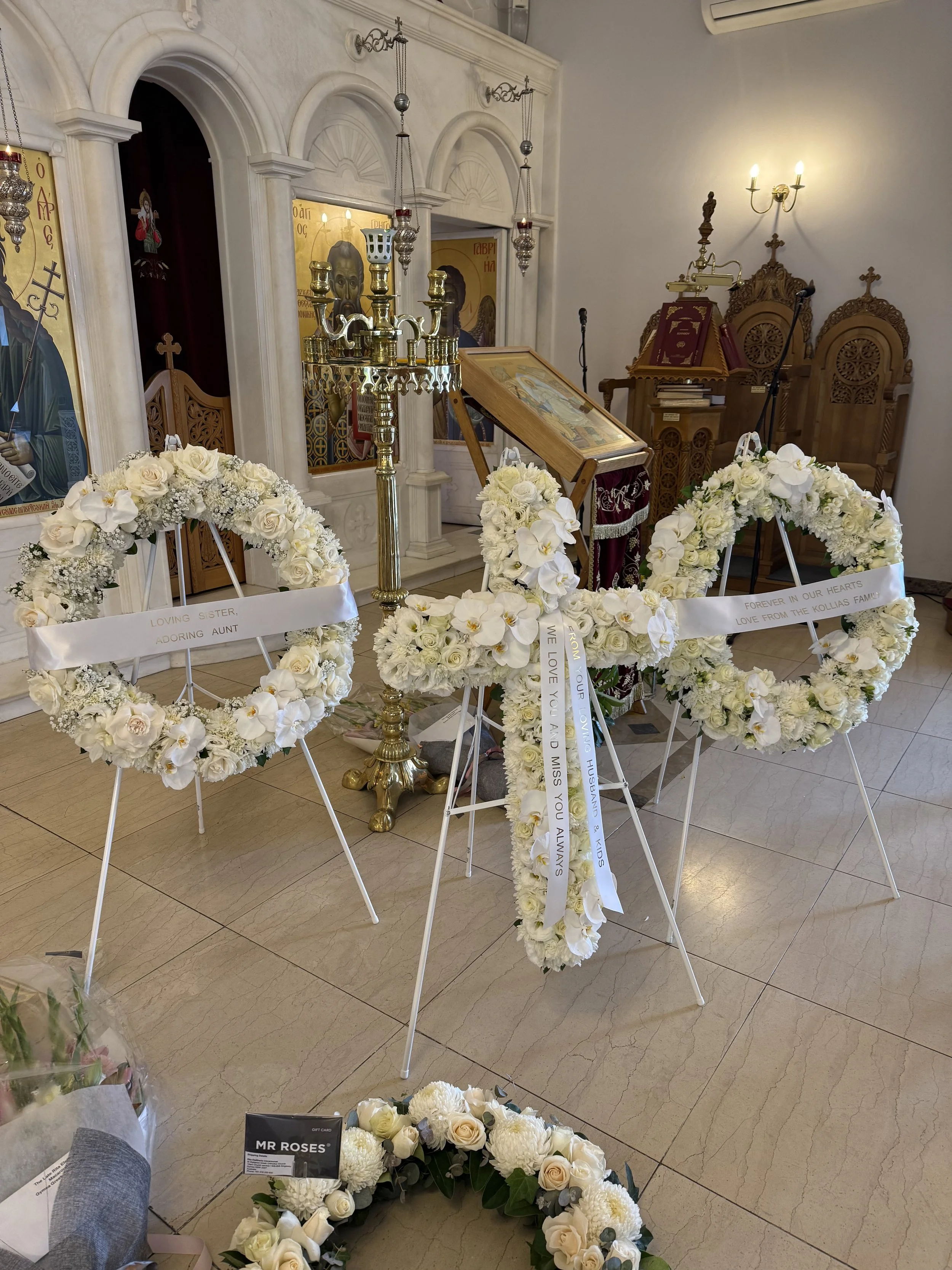 Funeral altar with white floral wreaths, icons, candles, and a small speaker in a church interior.
