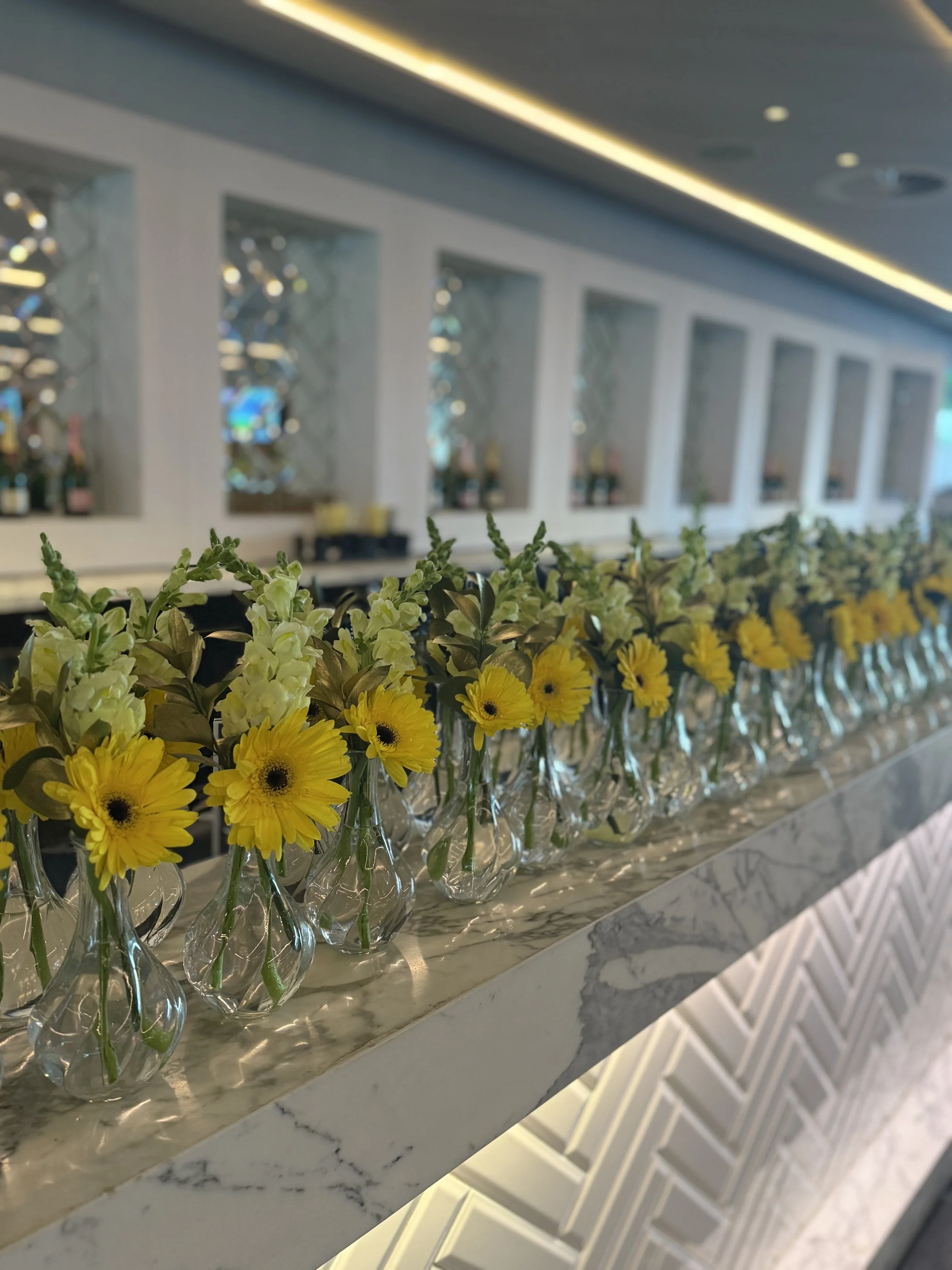 A row of glass vases with yellow and green flowers on a marble countertop, set against a background of decorative wall panels and mirrors.