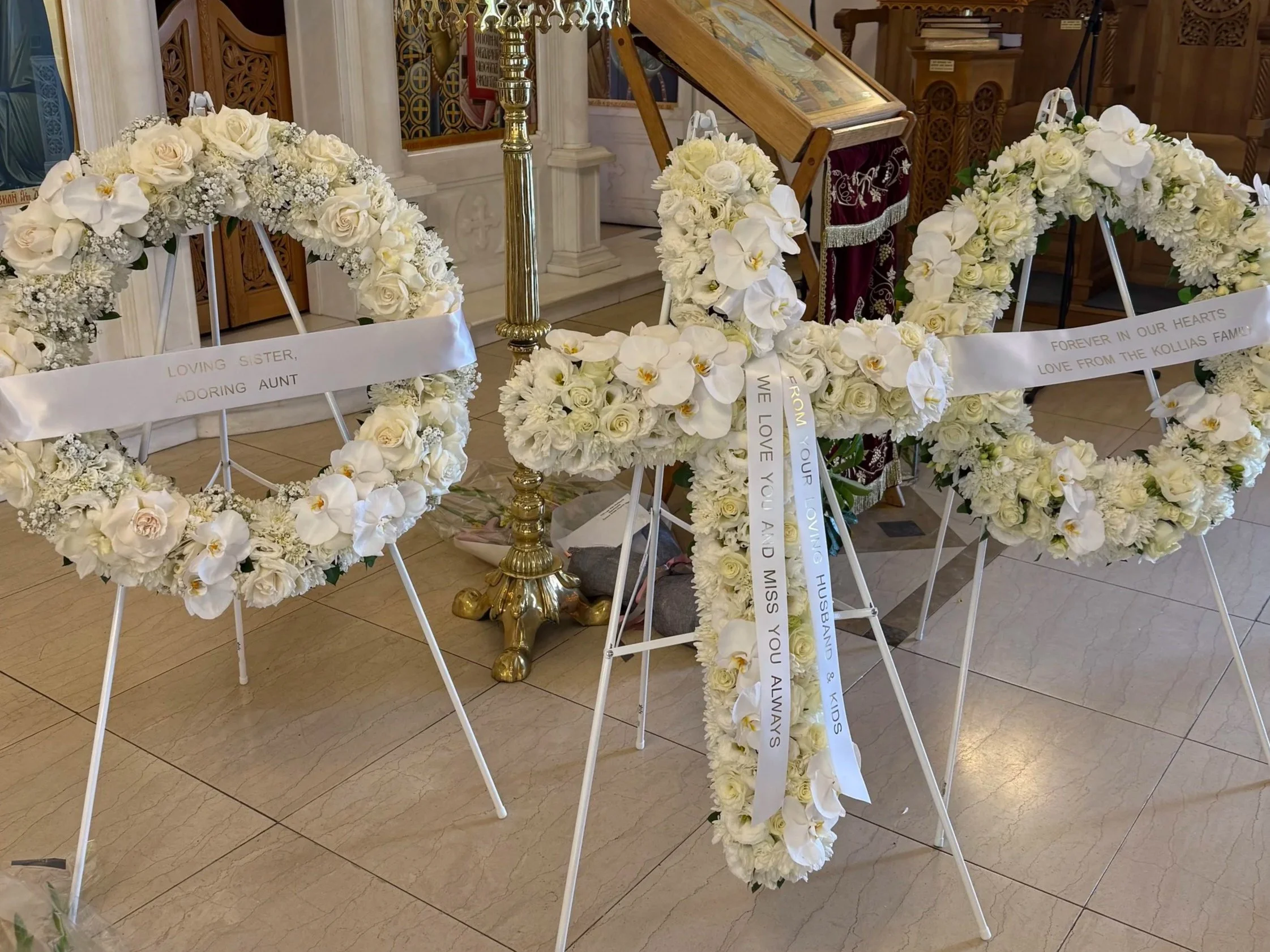 A display of white floral wreaths and arrangements arranged on metal stands inside a church or chapel, with messaging ribbons attached to some of the wreaths.