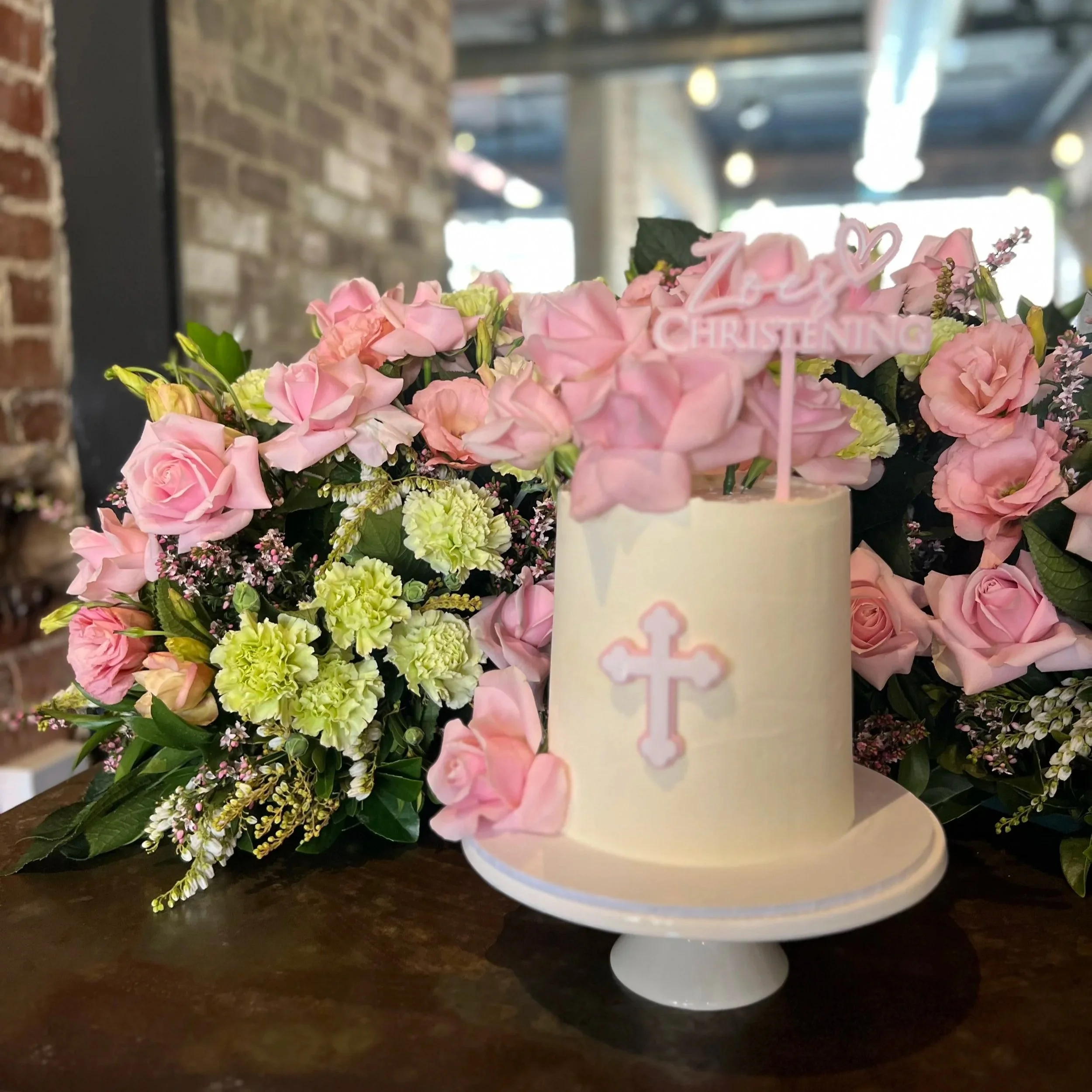A floral arrangement on a dark wooden surface, featuring pink roses, light pink hydrangeas, and small lavender flowers, with a pink sign that reads 'Baby Christening.'