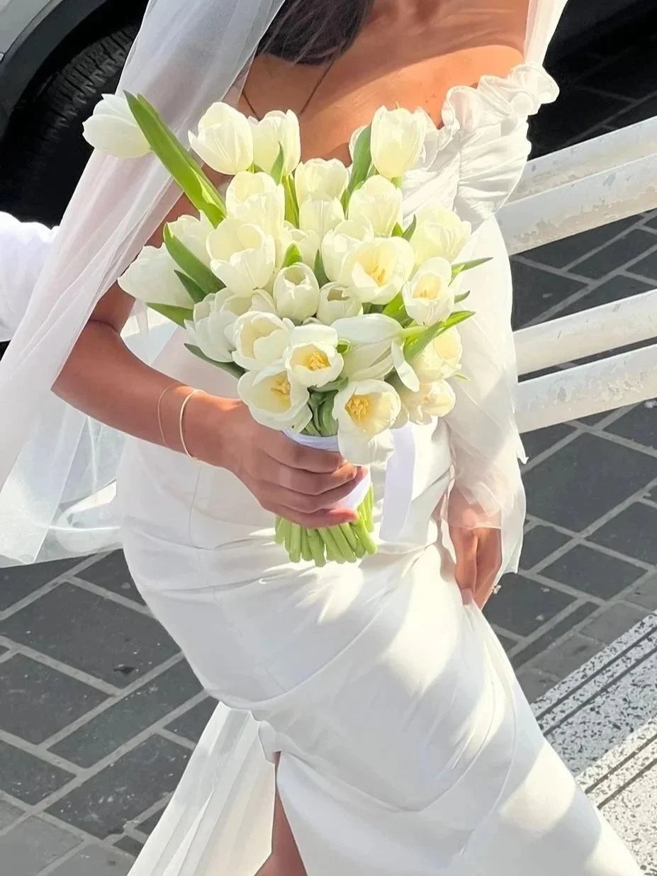 A woman in a white bride gown holding a bouquet of white tulips, sitting on a white bench outdoors.