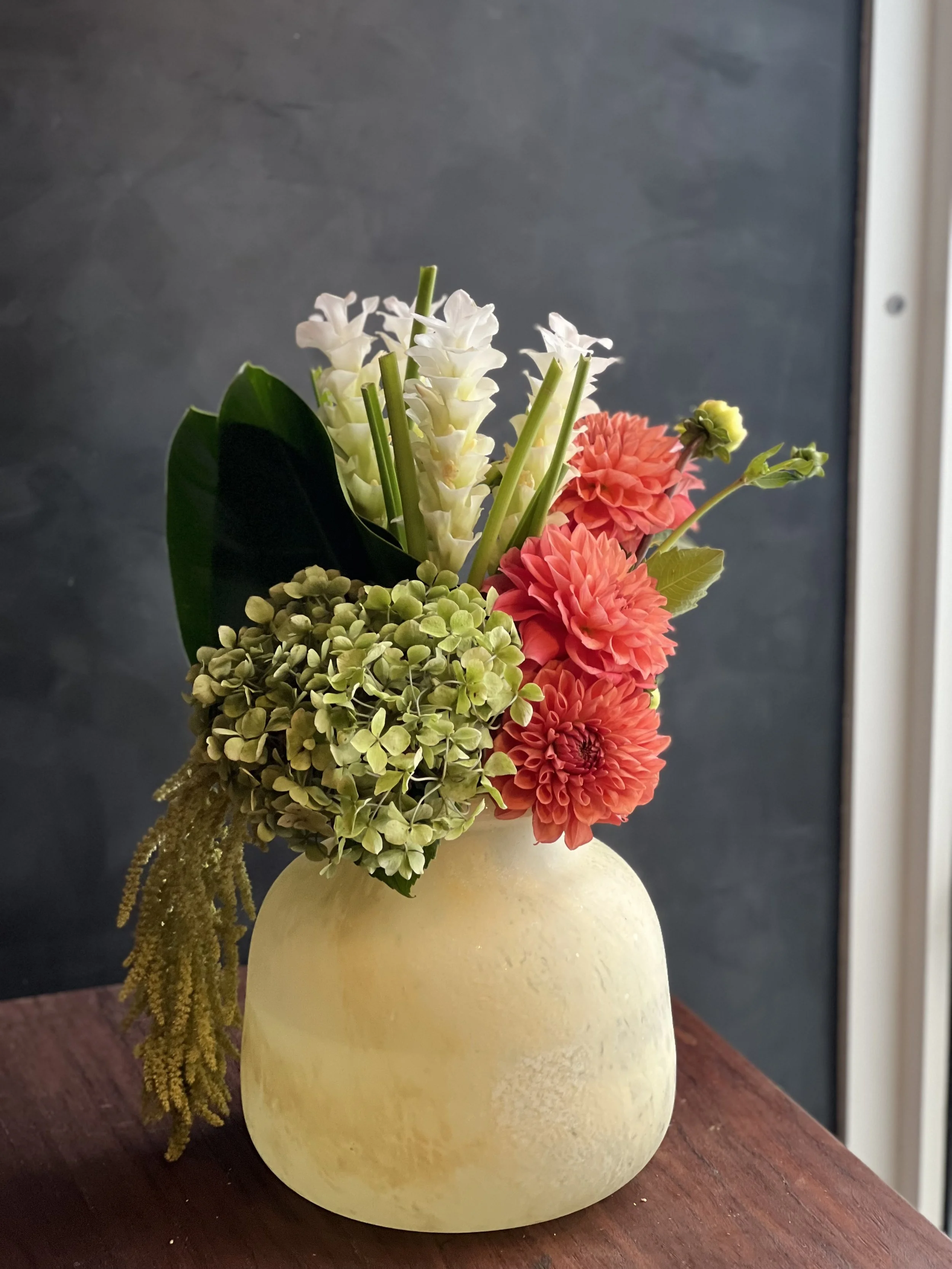 A floral arrangement in a cream-colored, round vase containing white, pink, green flowers, and greenery on a wooden surface.