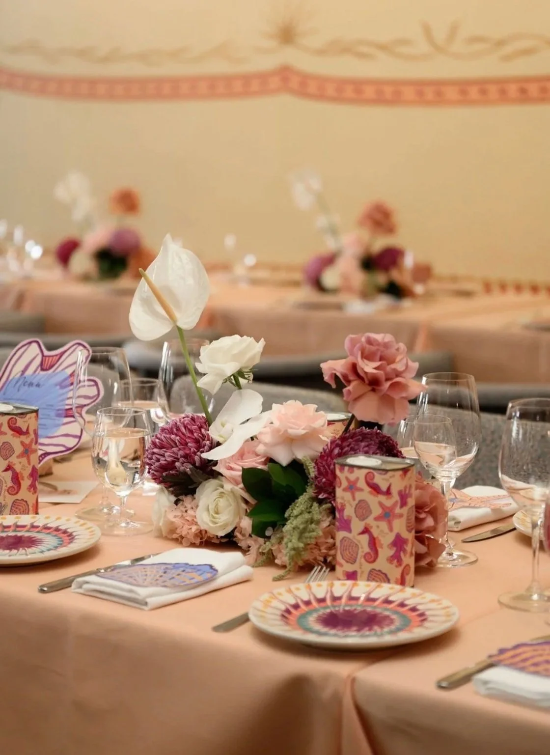 A decorated banquet table set with floral centerpieces, colorful plates, wine glasses, and napkins in a formal event room featuring pink and beige tones and hanging wall decorations.