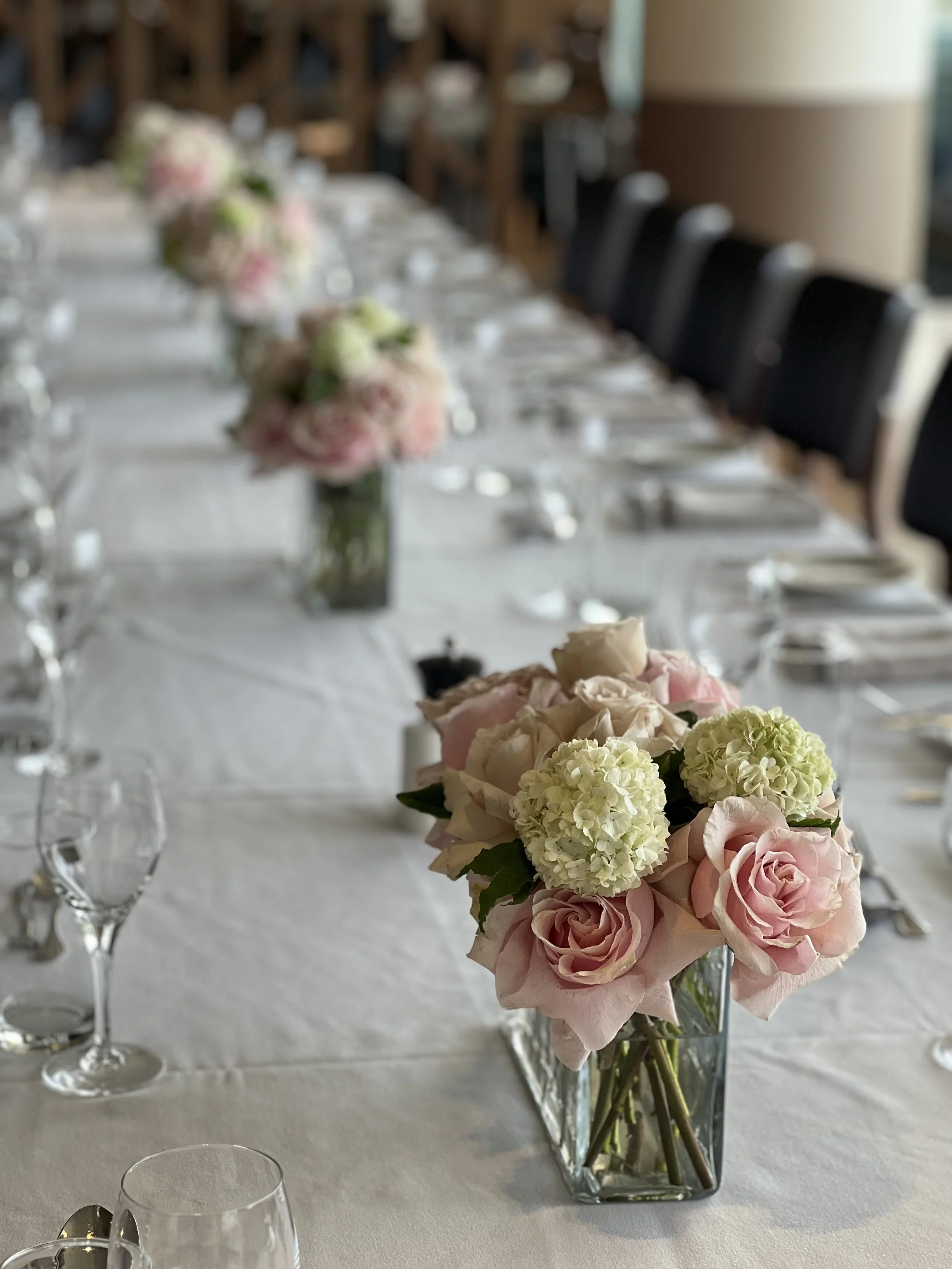 A table setting with floral centerpieces consisting of pink roses, white hydrangeas, and light green flowers in square glass vases, along with empty wine glasses and cutlery on a white tablecloth at a formal event.