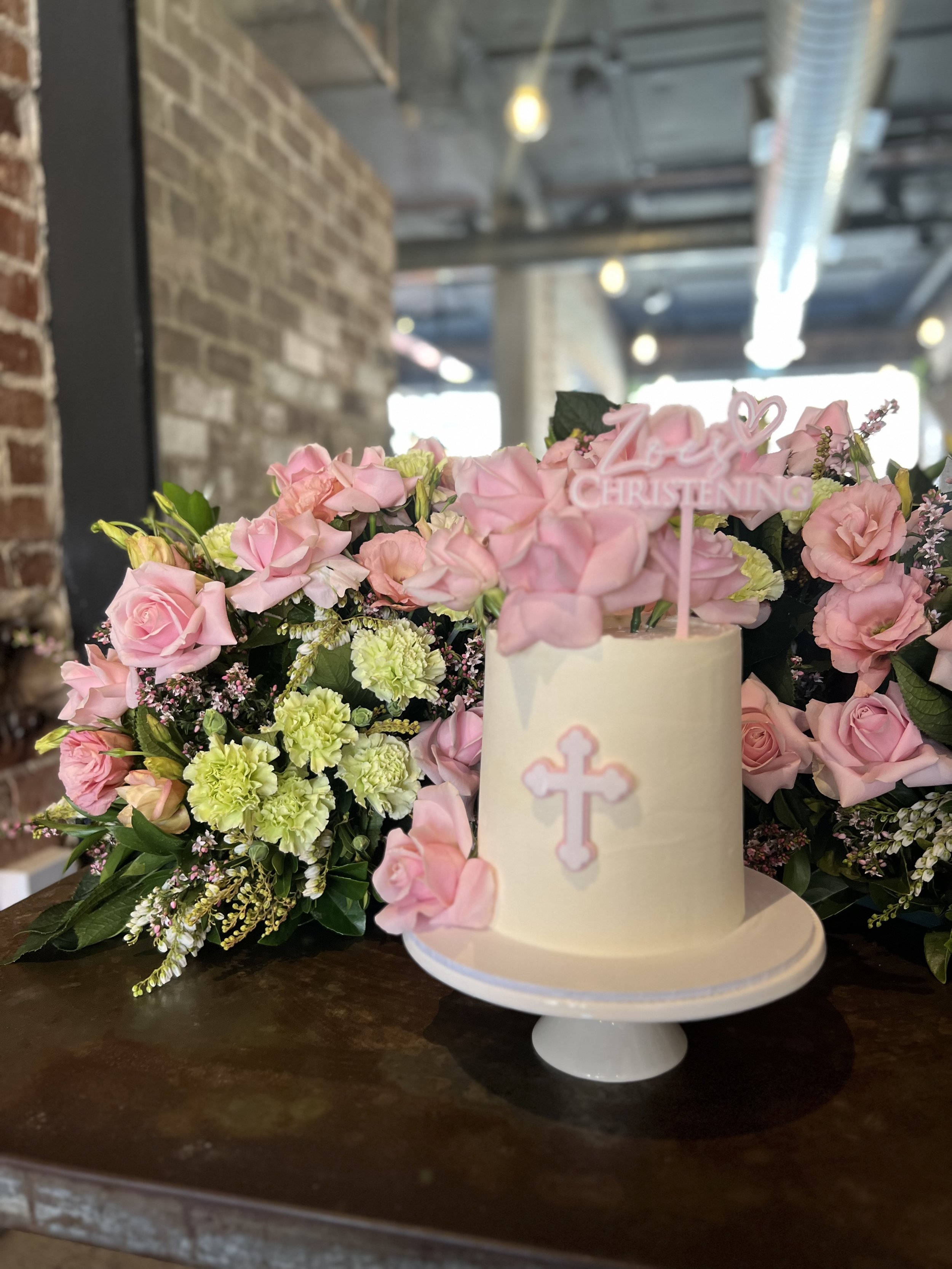 A pink and white baptism cake with a cross and a sign that says 'Love Christening' is surrounded by pink roses and green foliage on a dark wooden table in an industrial-style setting with exposed brick walls and ceiling pipes.