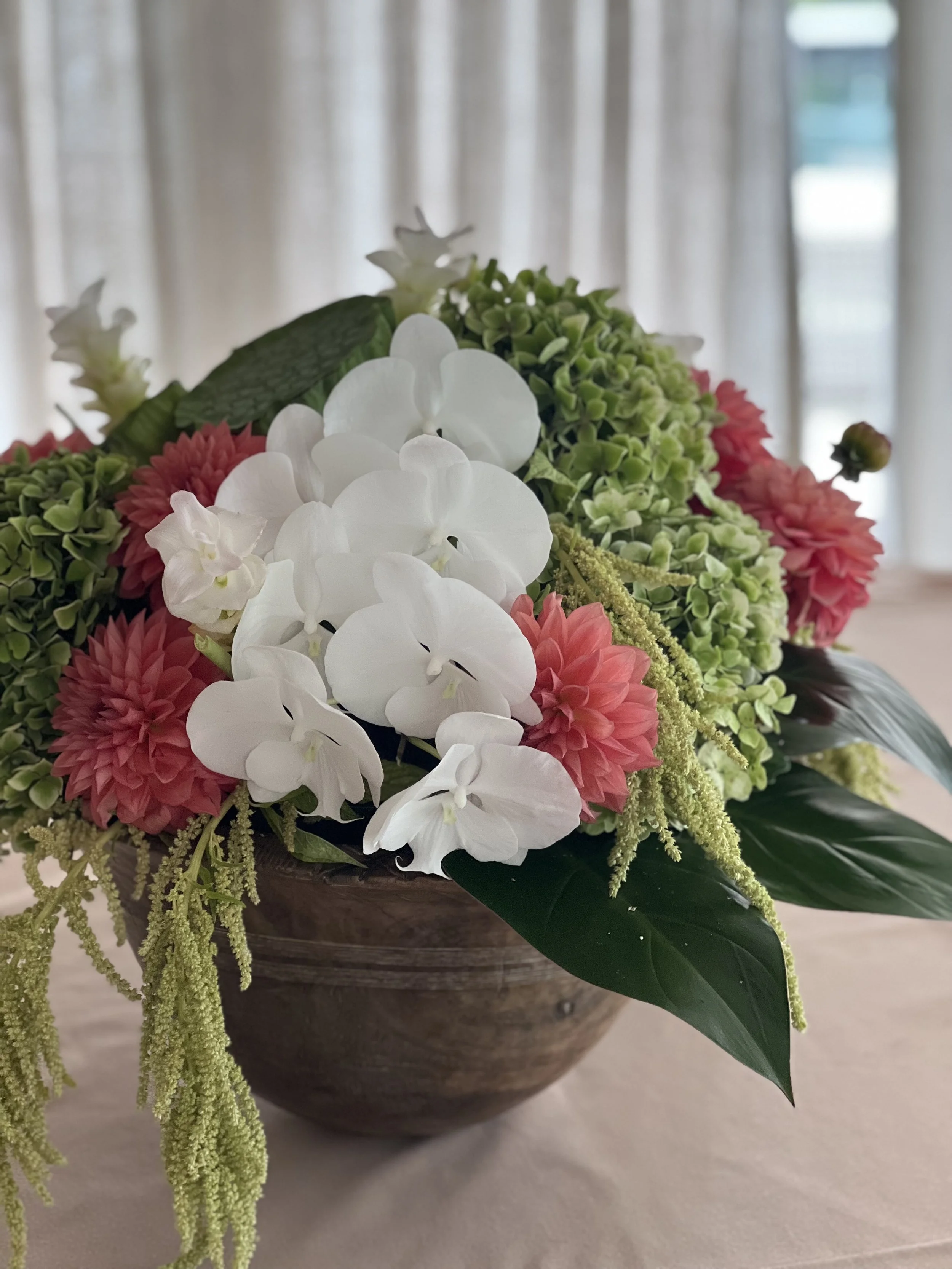 A floral arrangement in a wooden bowl featuring white, pink, and green flowers with large green leaves, set against a background of white curtains.