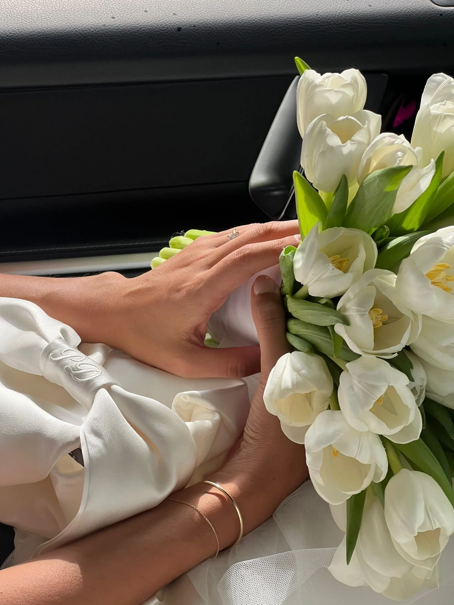 Close-up of a bride and groom holding a bouquet of white flowers on a wedding day, with sunlight illuminating their hands and the flowers.