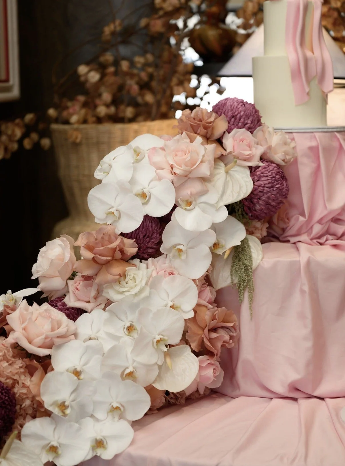 Close-up of a decorative floral arrangement with white orchids, pink roses, and purple flowers on a pink fabric-covered table, with a basket of dried flowers and a lamp in the background.