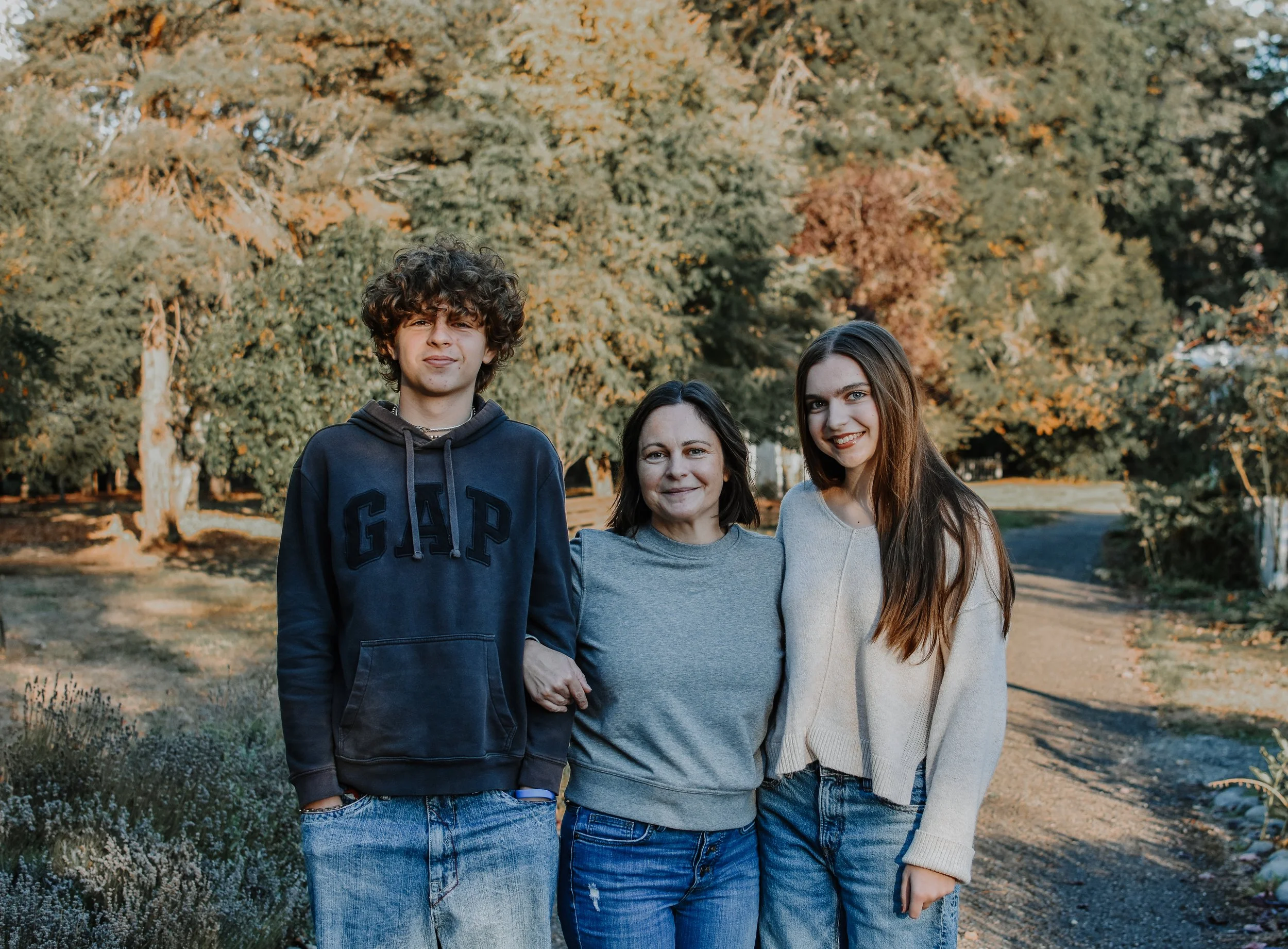 Three people standing together outdoors during fall, with trees and a path in the background.