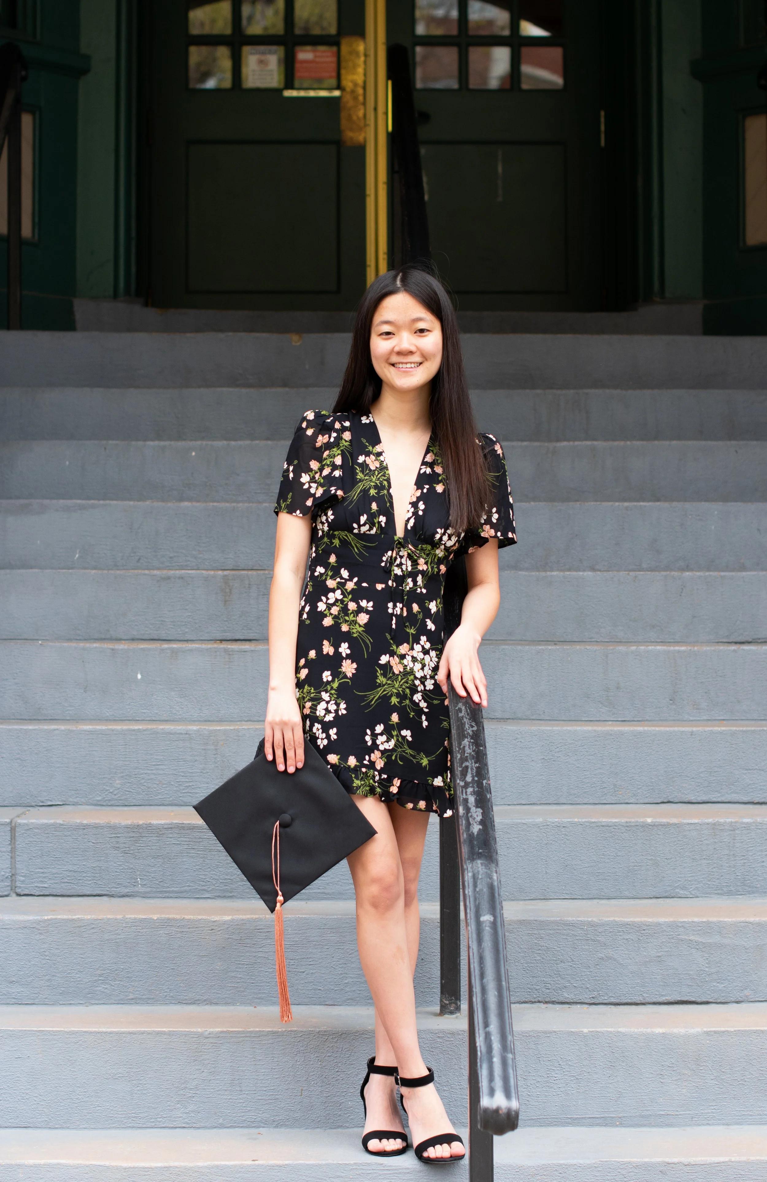 Young woman in a floral dress holding a graduation cap, standing on stairs outside a building.