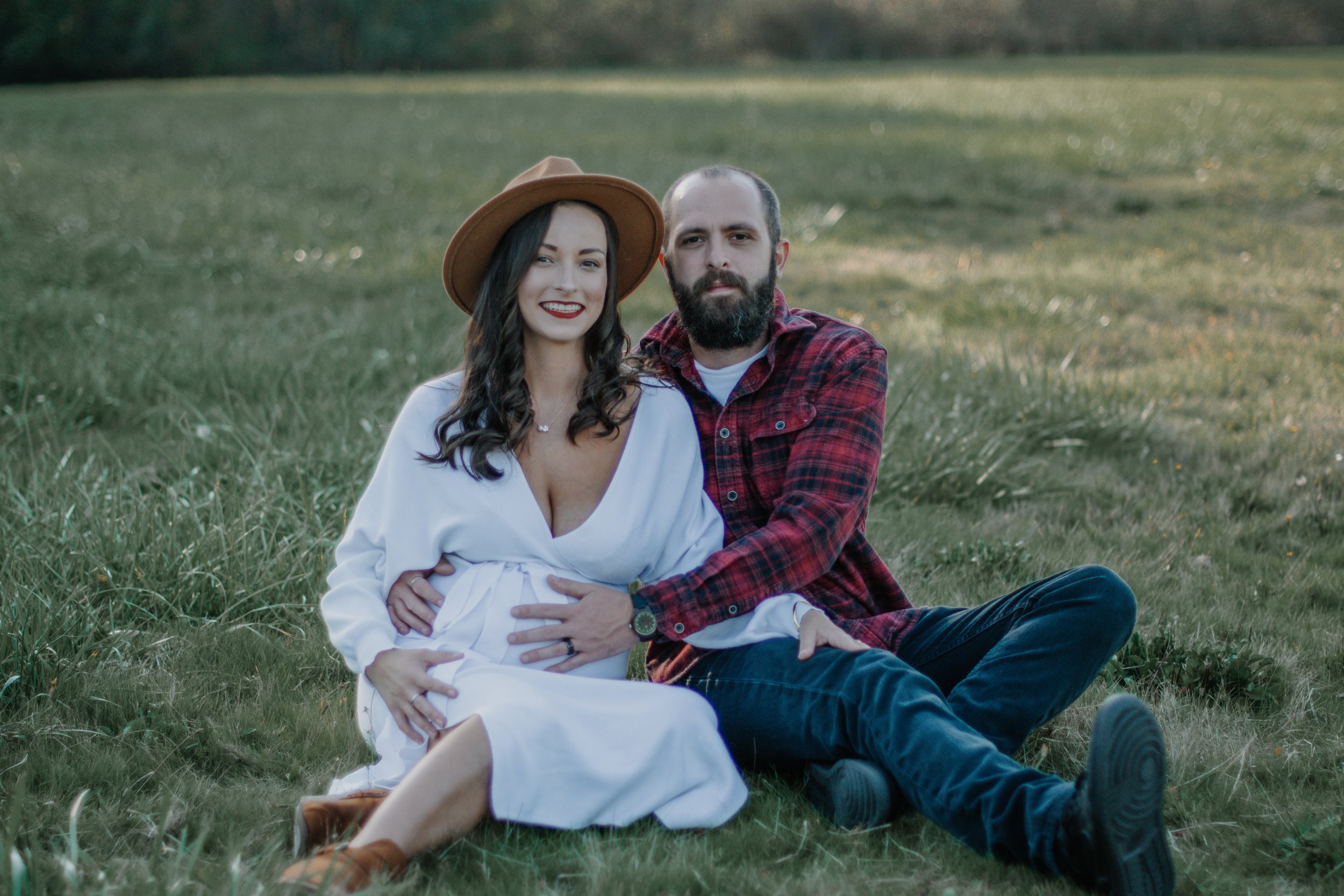 A couple sitting on the grass in a field, with the woman wearing a white dress and hat, and the man wearing a red plaid shirt, both smiling at the camera.