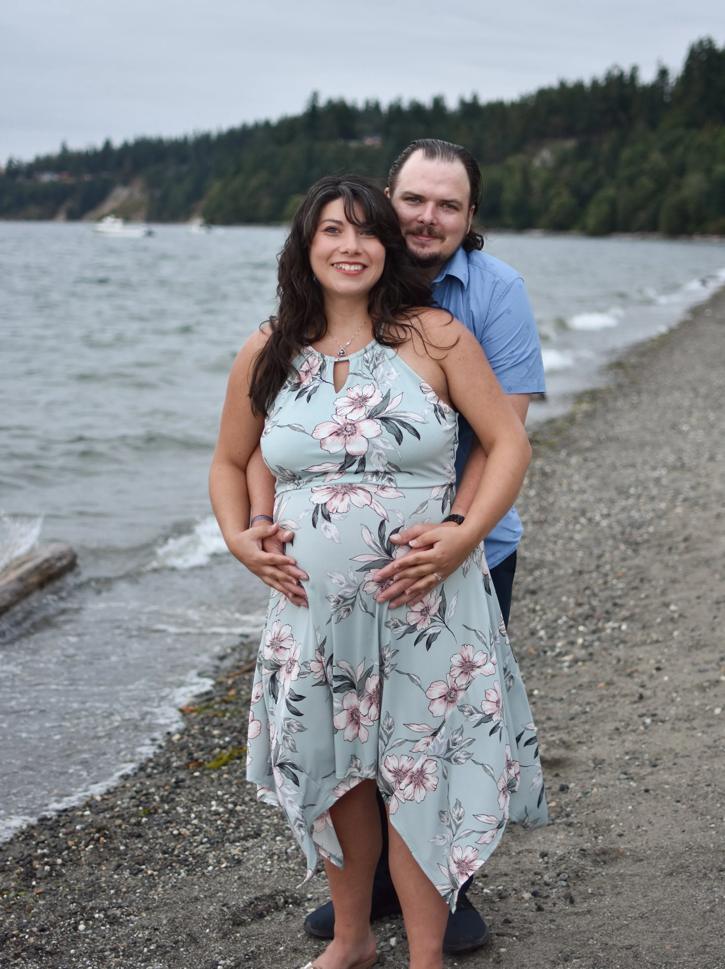 A pregnant woman and a man standing on a beach by the water, with the woman in front and the man standing behind her with his arms around her waist. The woman is smiling and wearing a floral dress, while the man looks at the camera and is dressed in 