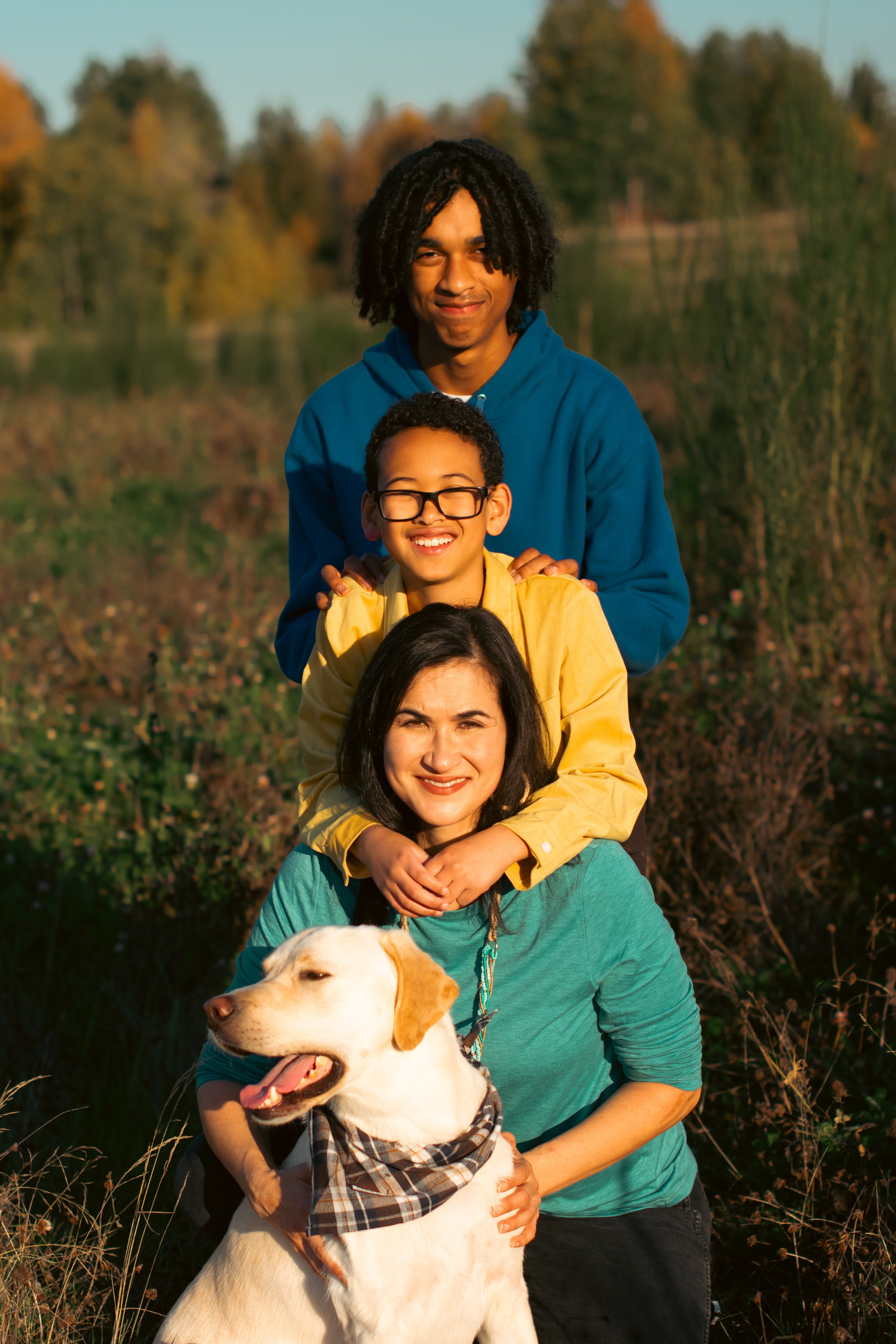A family of four, including a dog, posing outdoors in a line, smiling. The background features trees with fall foliage.