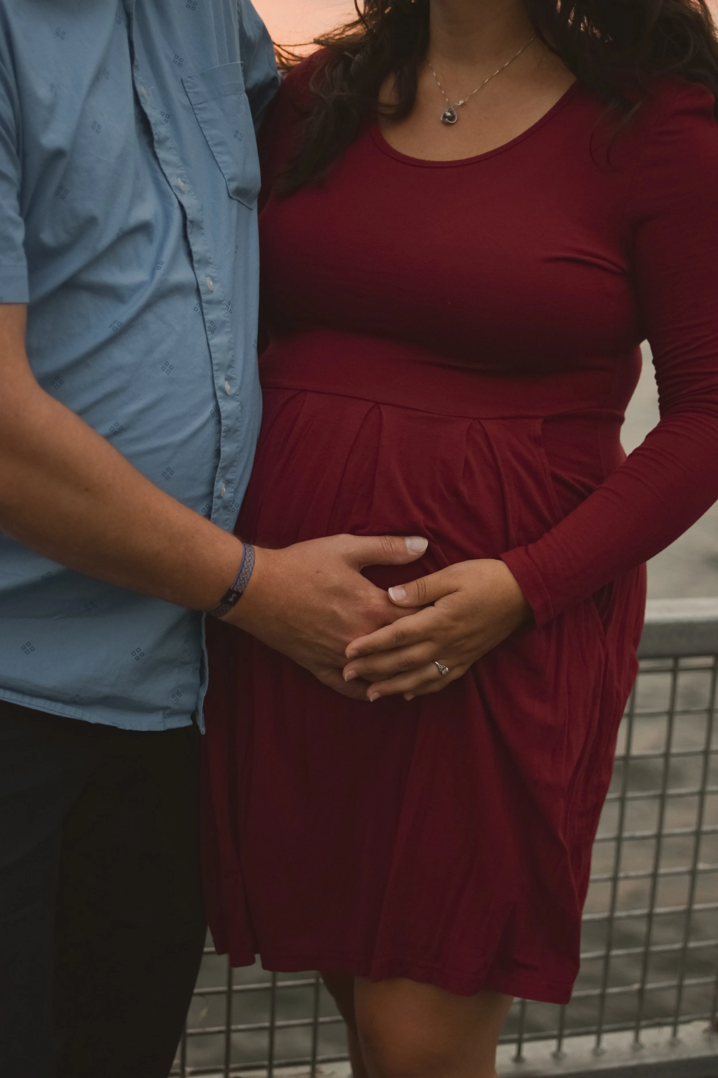 A pregnant woman wearing a red dress holding her belly with her hands, standing next to a man in a blue shirt with his hand on her belly, on a bridge during sunset.