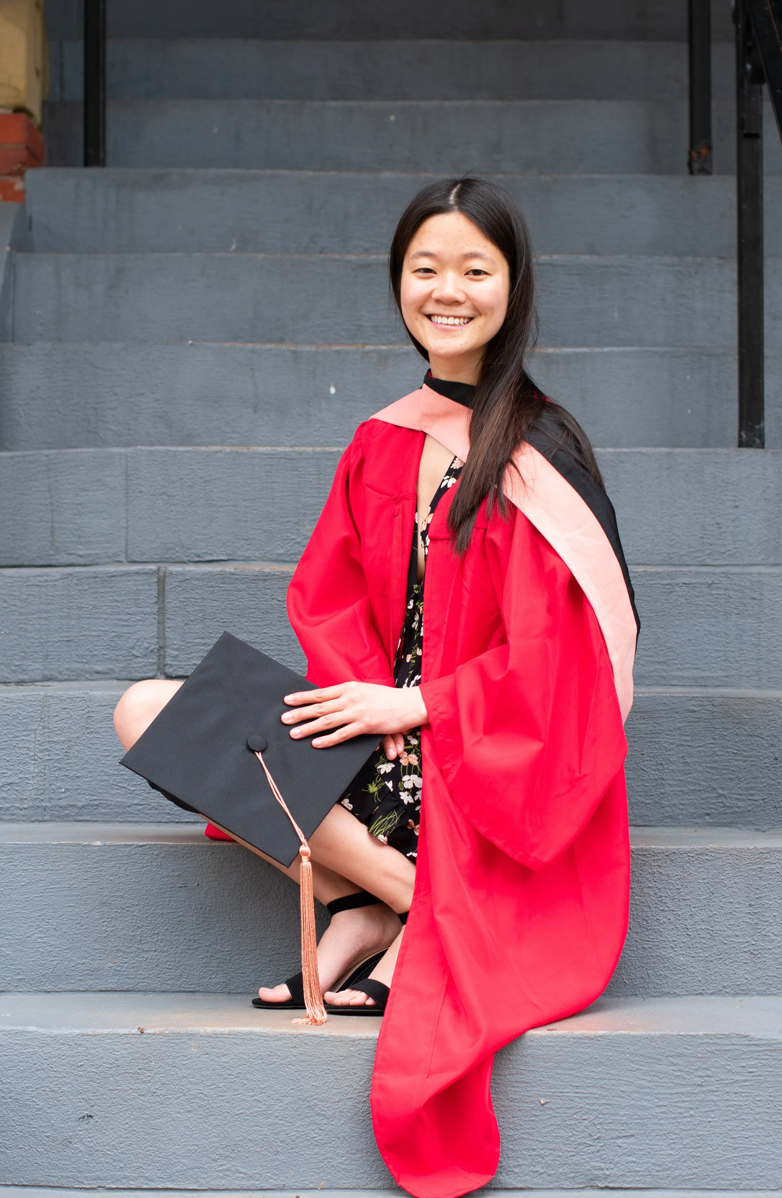 A young woman in a red graduation gown and cap, holding a diploma, sitting on gray stairs and smiling.