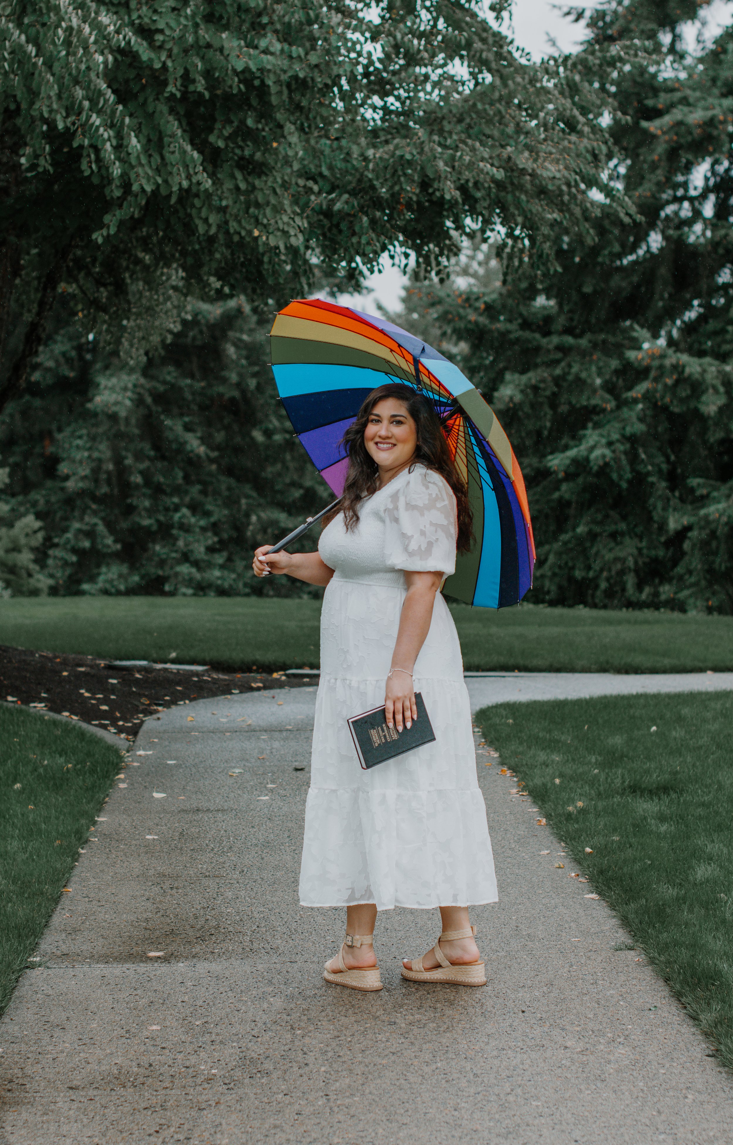 A woman in a white dress holding a colorful rainbow umbrella and a black book, standing on a park sidewalk with green trees in the background