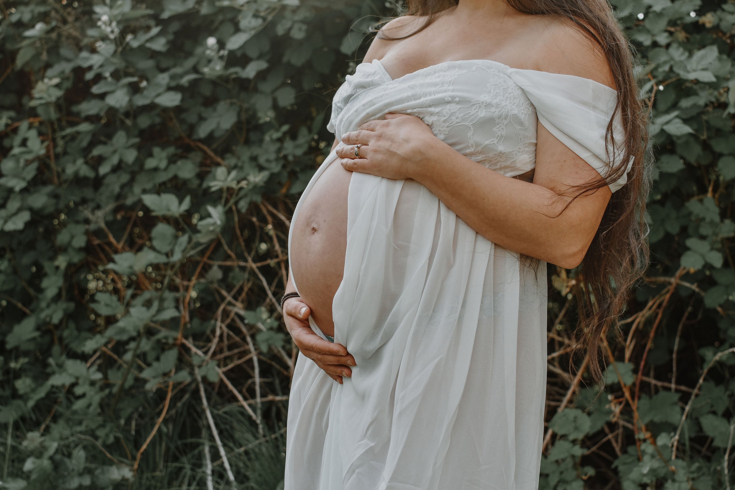 Pregnant woman in a white dress stands outdoors with green foliage in the background, holding her belly with both hands.