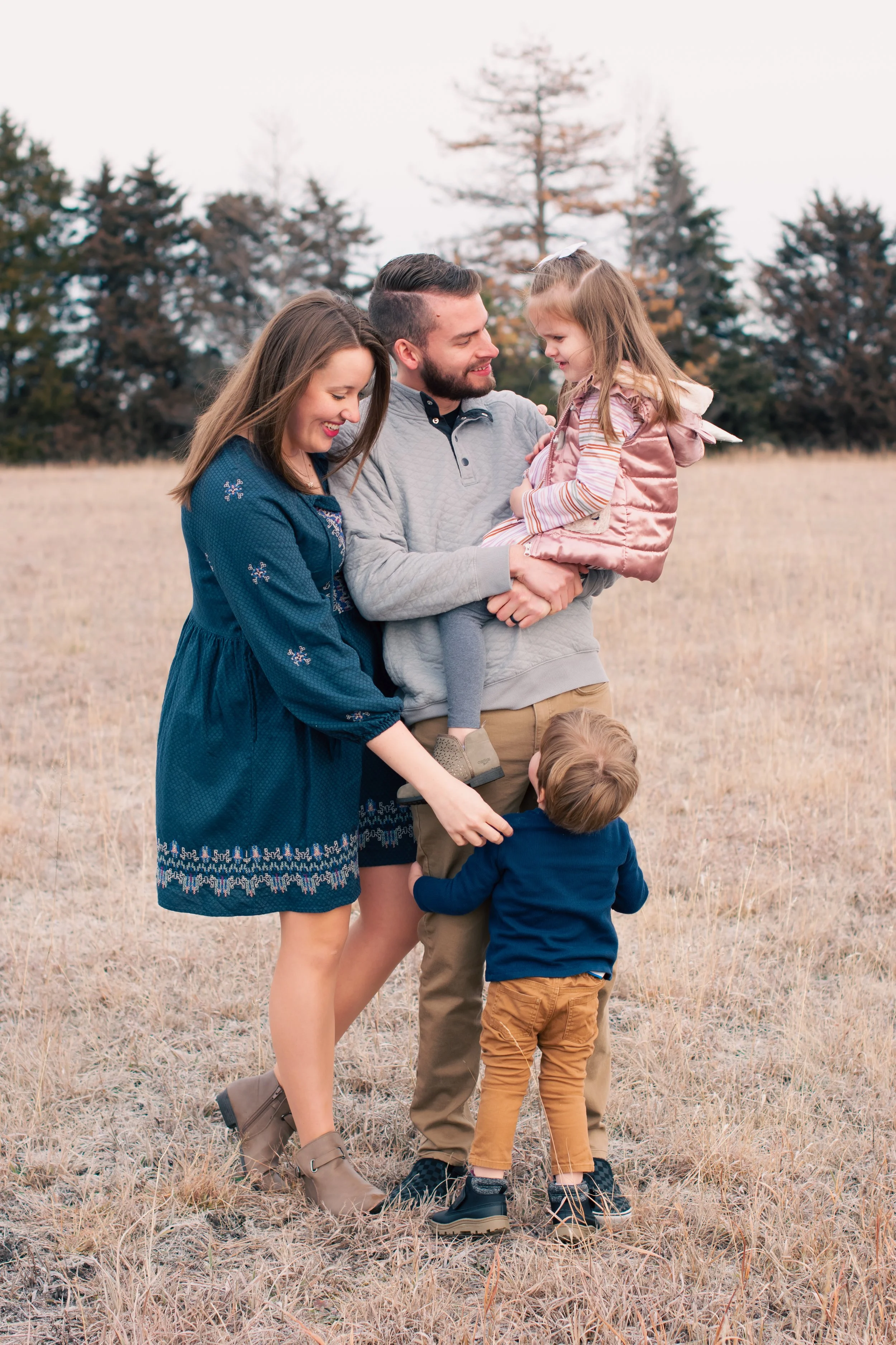 Family of four enjoying time outdoors in a field with trees in the background, a mother, father, and two young children smiling and playing together.