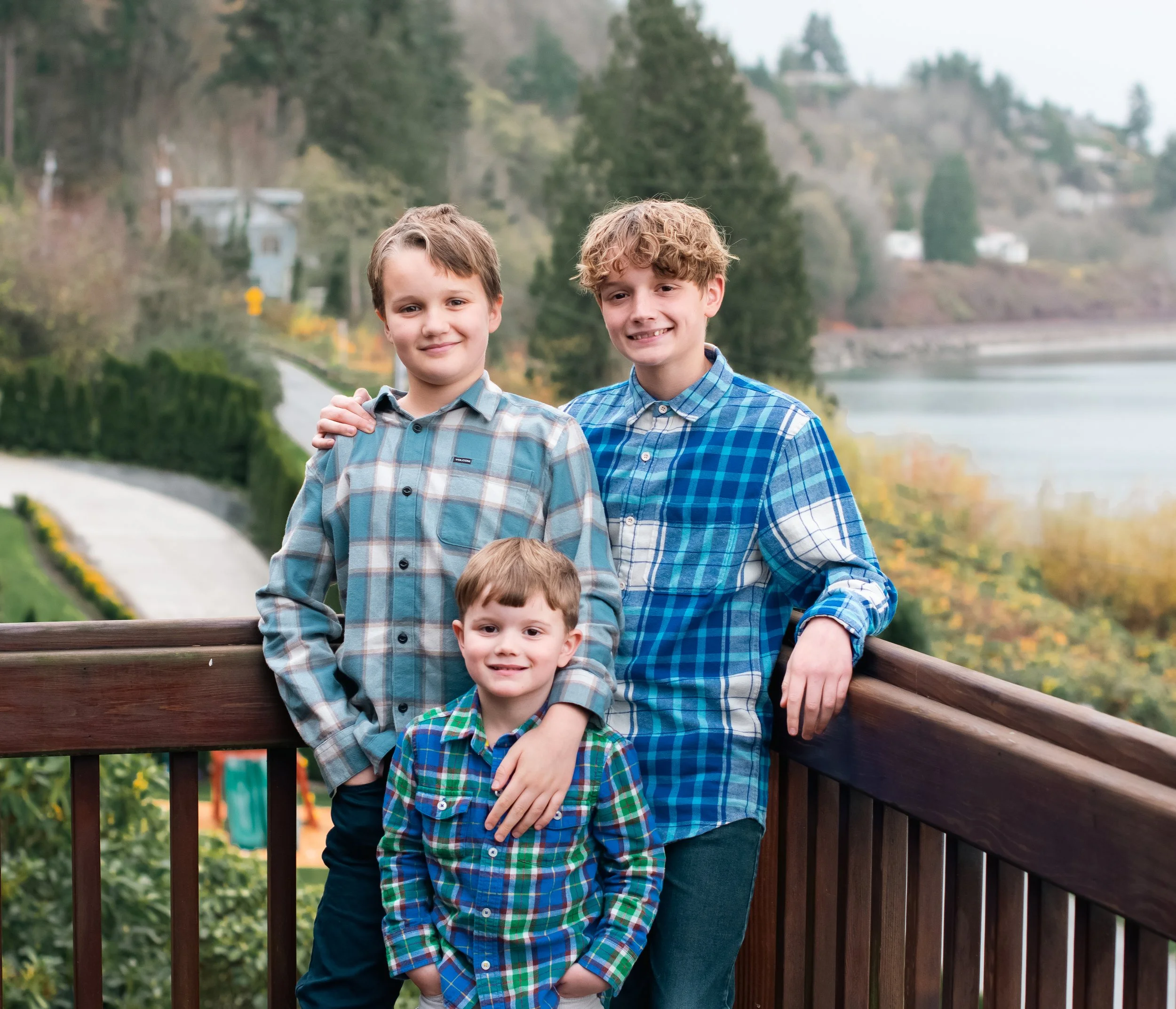 Three boys standing on a wooden deck with a scenic lake and trees in the background. The oldest has curly hair and is wearing a blue plaid shirt, the middle boy has straight hair and is wearing a gray plaid shirt, and the youngest has straight hair a