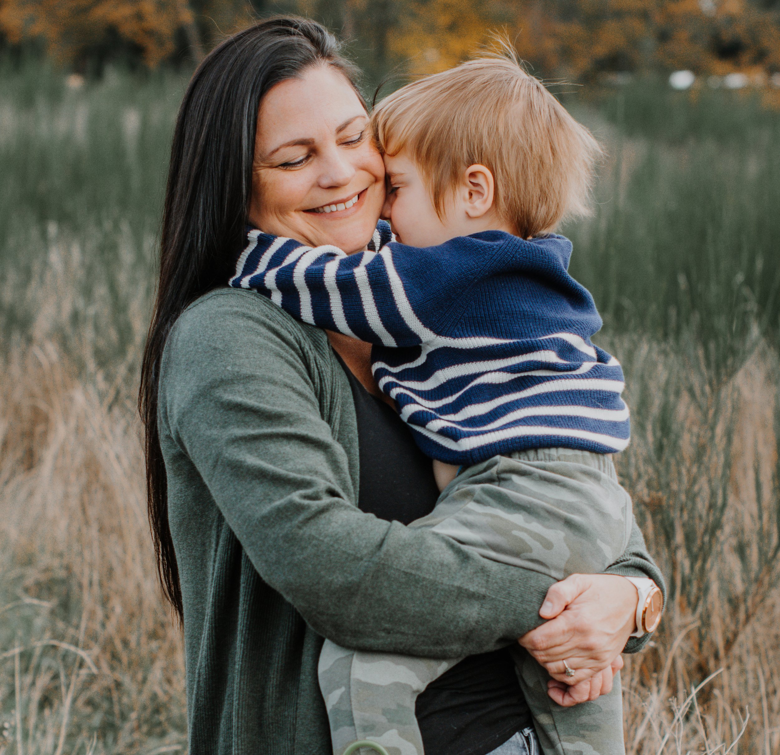 A woman holding a young boy outdoors, both smiling and embracing affectionately, with a grassy field and trees in the background.