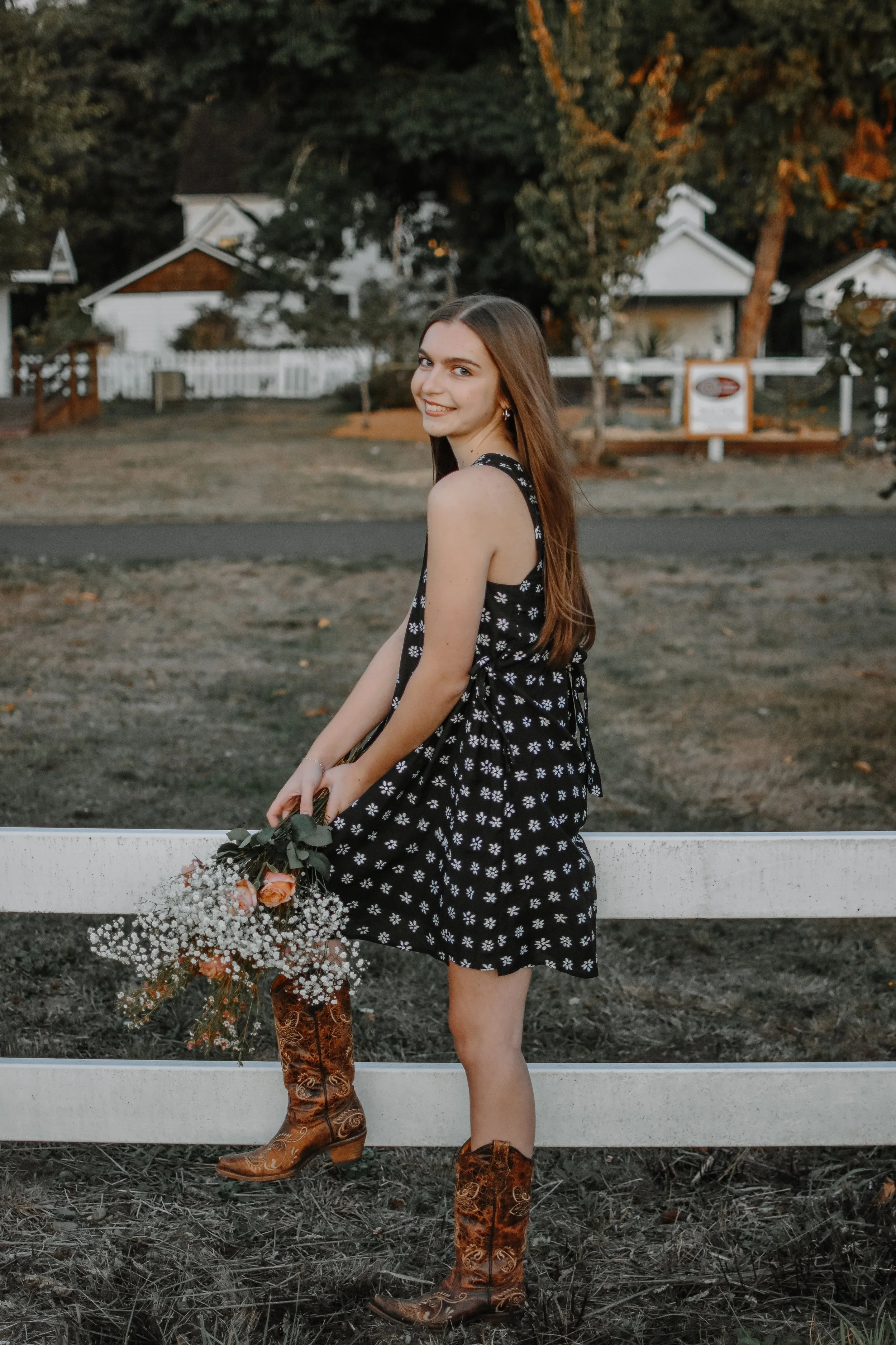 A young woman in a black dress with white floral pattern and cowboy boots, holding a bouquet of flowers, standing by a white fence in a rural setting with houses and trees in the background.