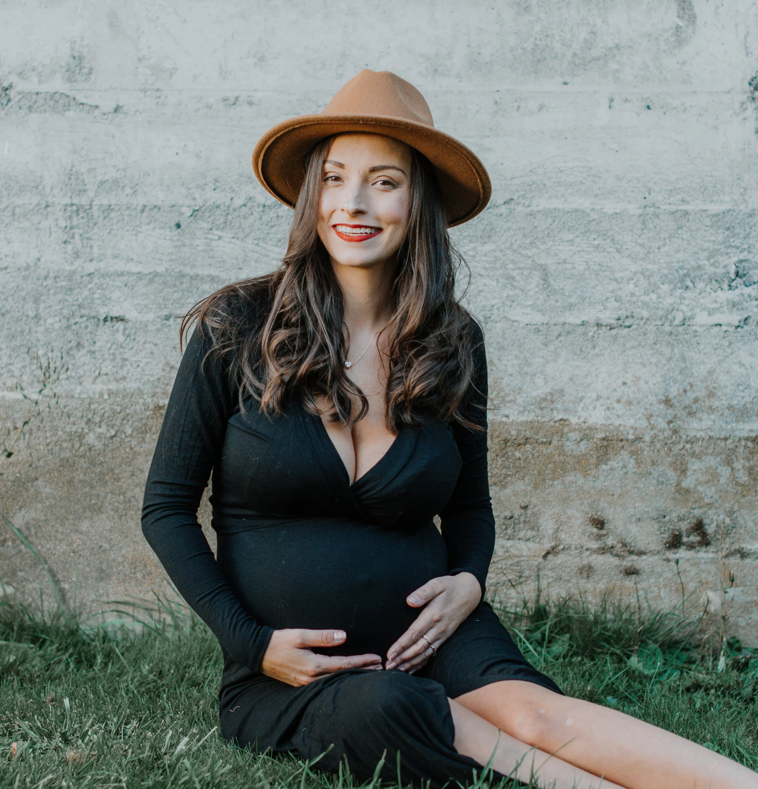 A smiling pregnant woman sitting on grass, touching her baby bump, wearing a black dress and a wide-brimmed brown hat, against a concrete wall background.