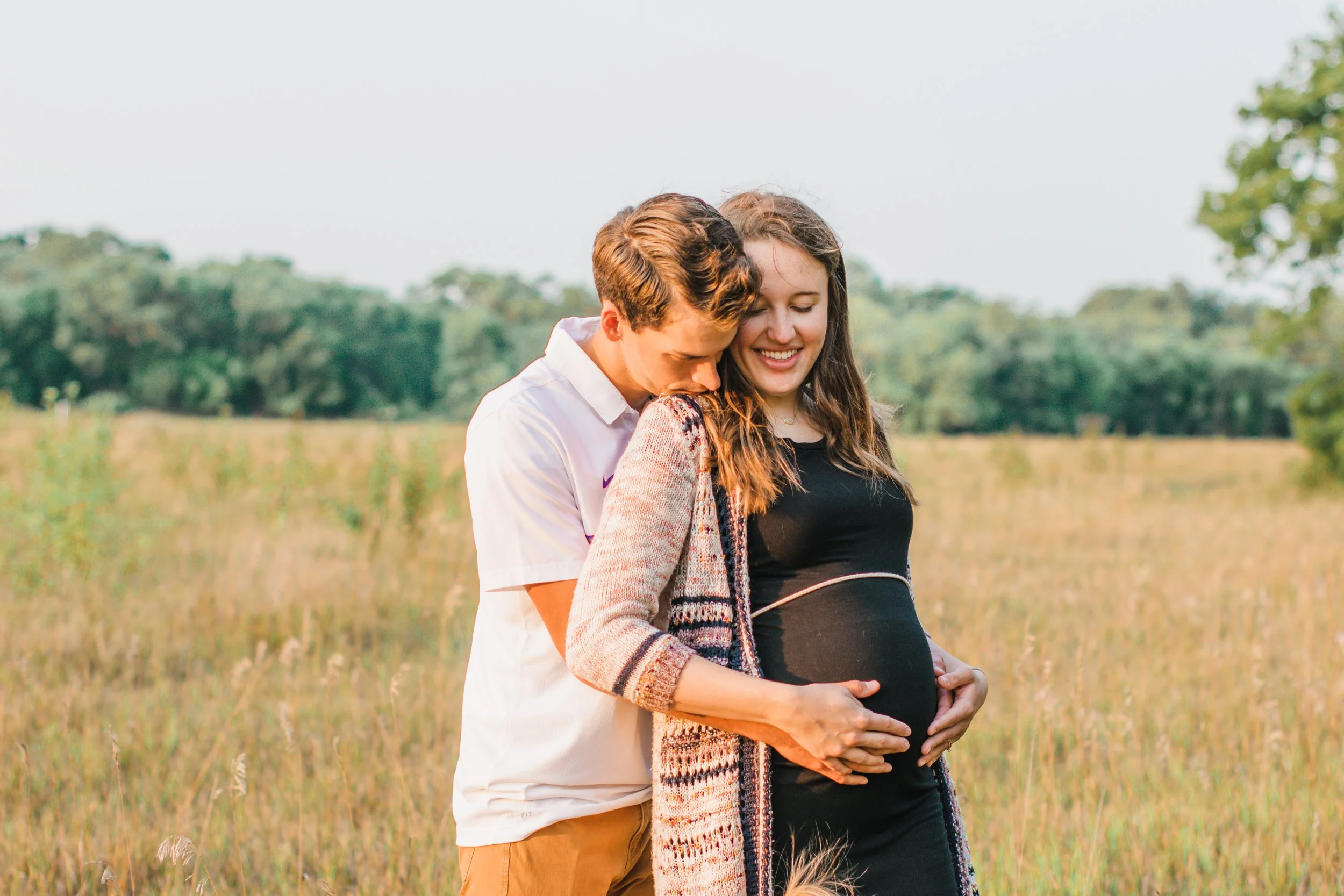 A young couple with a pregnant woman and her partner standing in a grassy field, smiling and touching her pregnant belly during sunset.