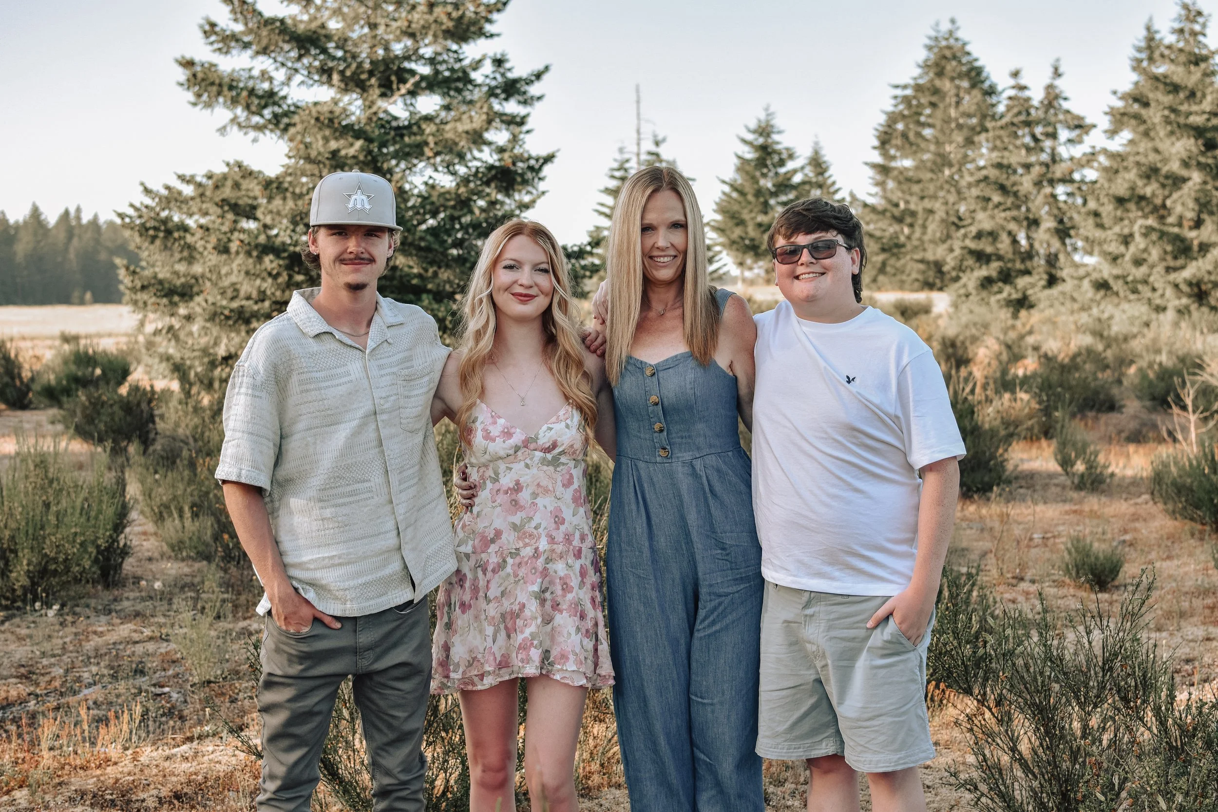 A group of four people, two men and two women, standing outdoors in a field with trees in the background. They are smiling and posing together.