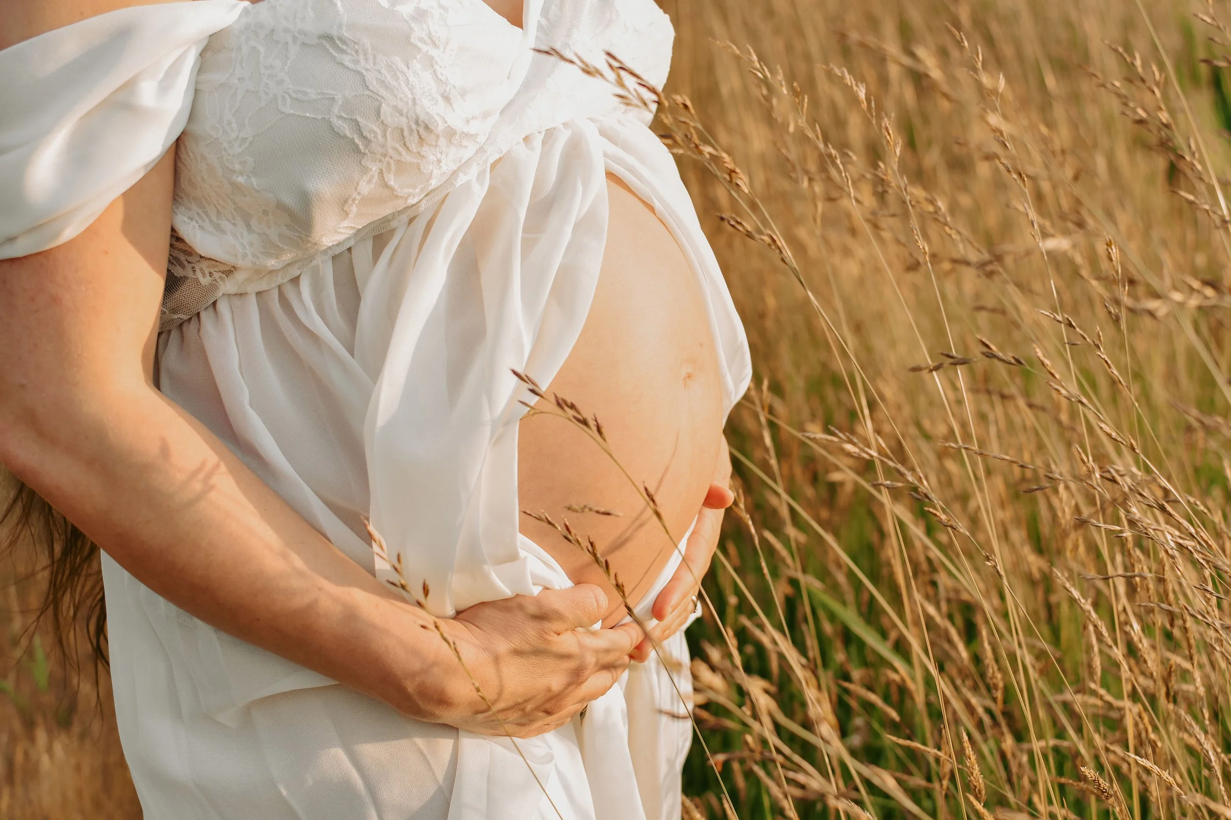 Close-up of a pregnant woman wearing a white dress, holding her belly with one hand, standing in a field of tall grass during sunset.