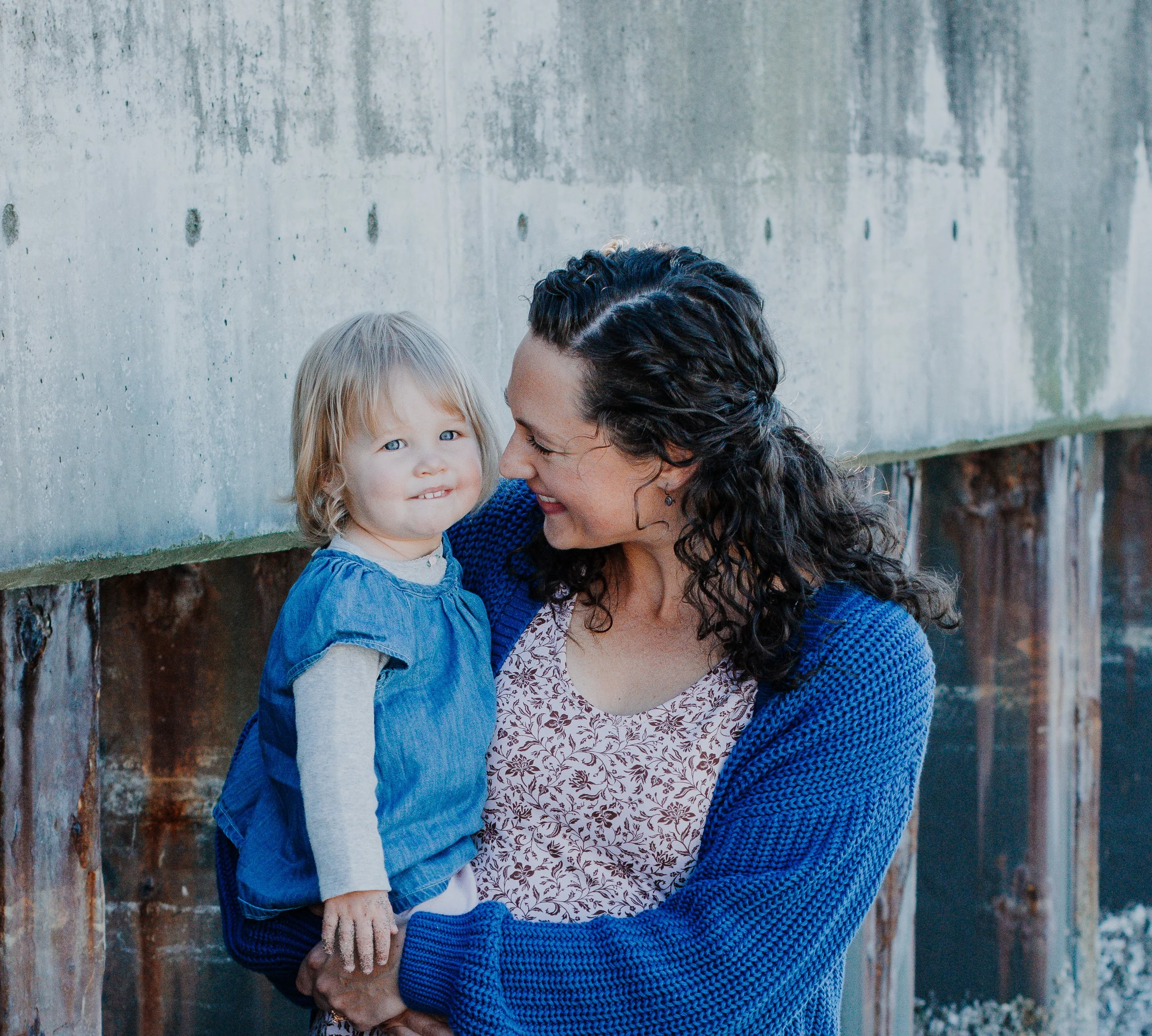 A woman with dark curly hair, wearing a blue cardigan, smiling and holding a young girl with blond hair, blue eyes, and wearing a blue dress and gray long sleeve shirt, standing outdoors near a weathered wooden and concrete wall.