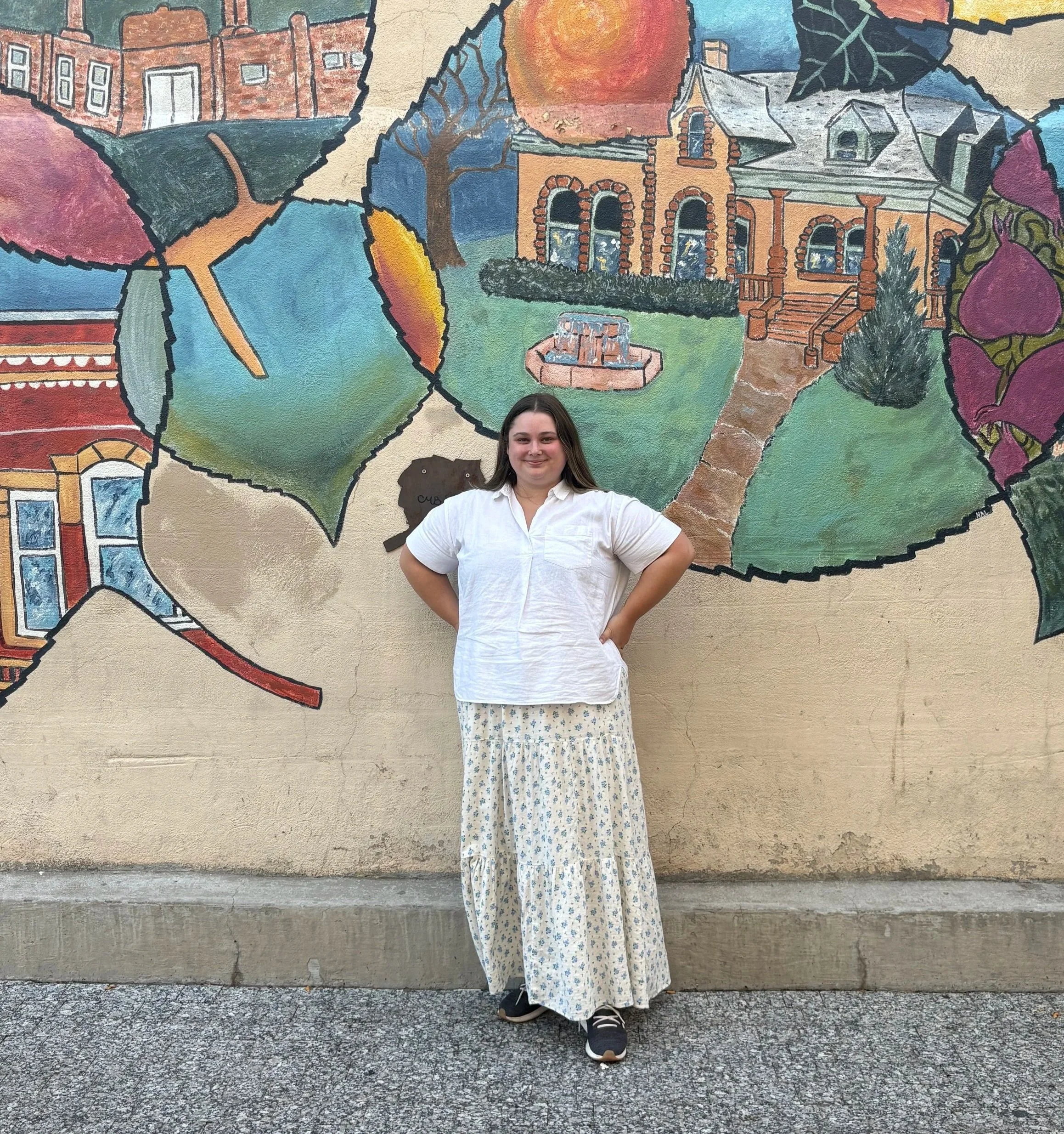 A woman standing in front of a colorful mural depicting various scenes and buildings, with her hands on hips, smiling at the camera.