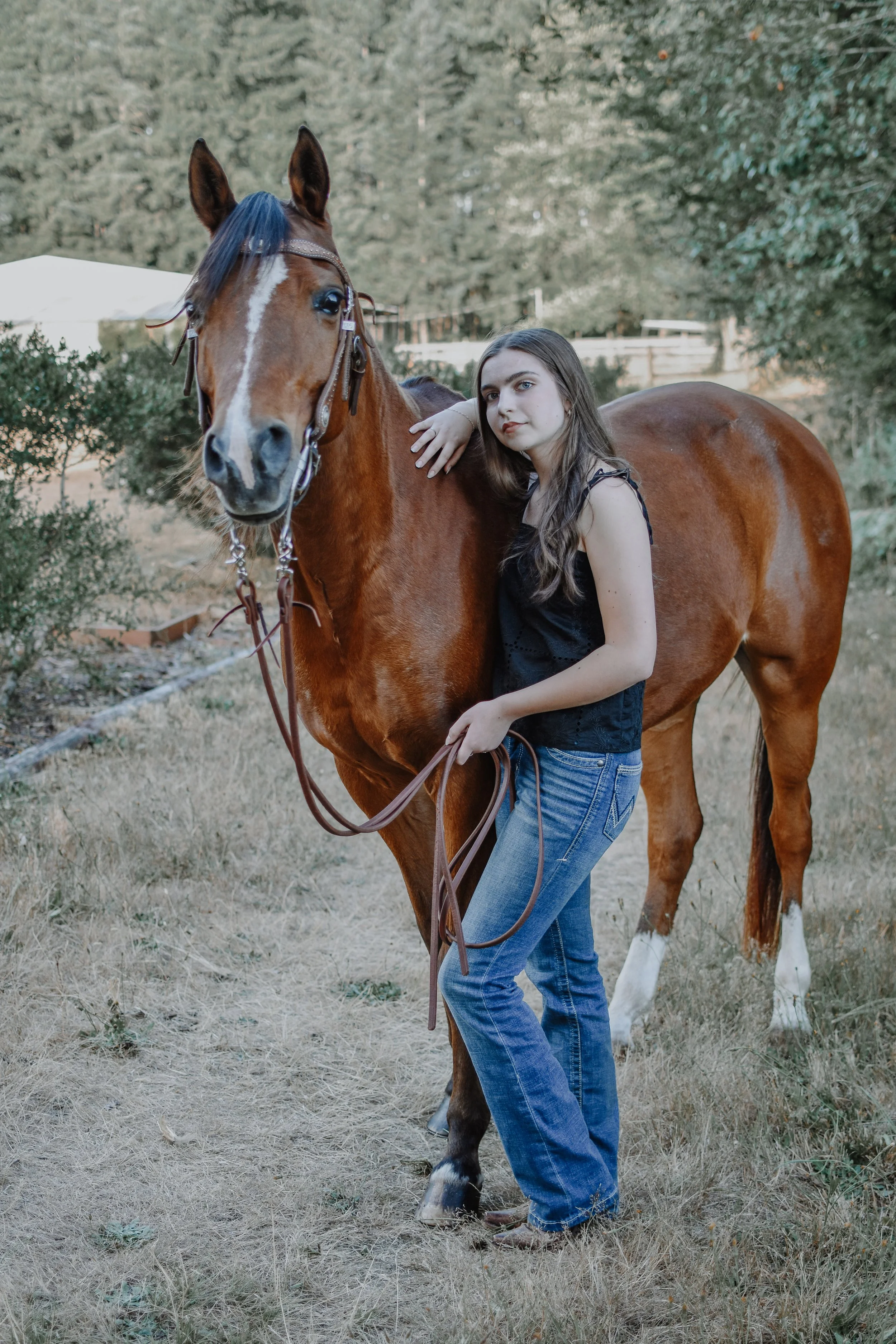 A young woman in jeans and a black sleeveless top standing next to a chestnut horse, holding its reins and resting her hand on its neck, in an outdoor grassy area with trees in the background.