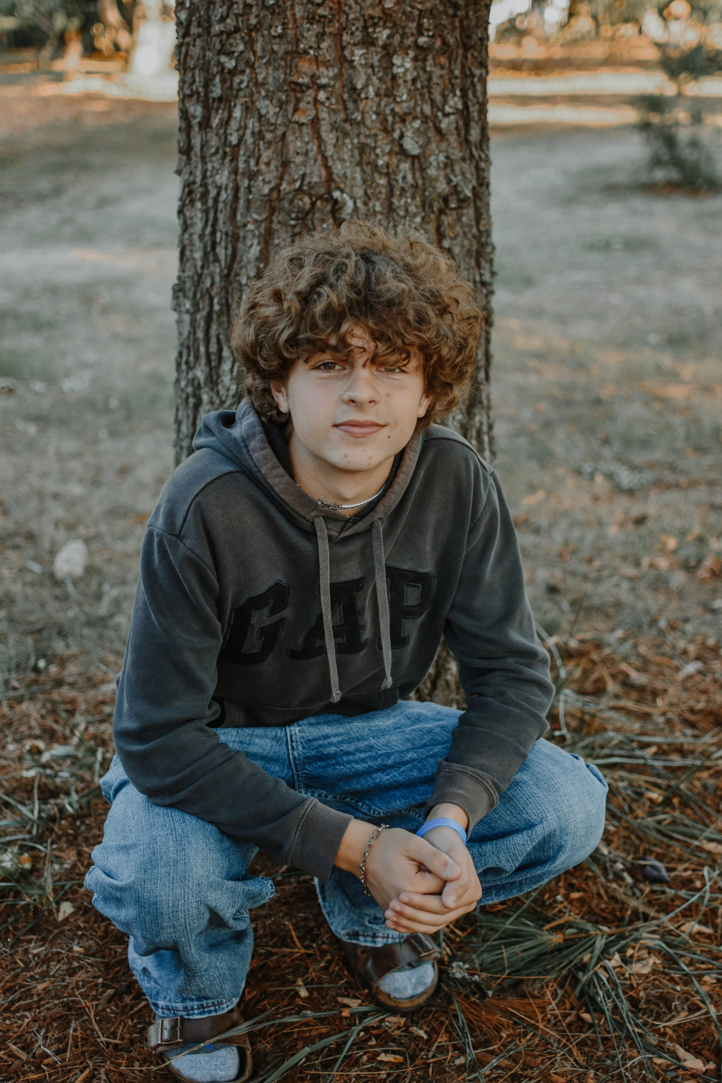 A young man with curly hair, wearing a gray hoodie and jeans, sitting on the ground with his arms crossed, in front of a large tree outdoors.