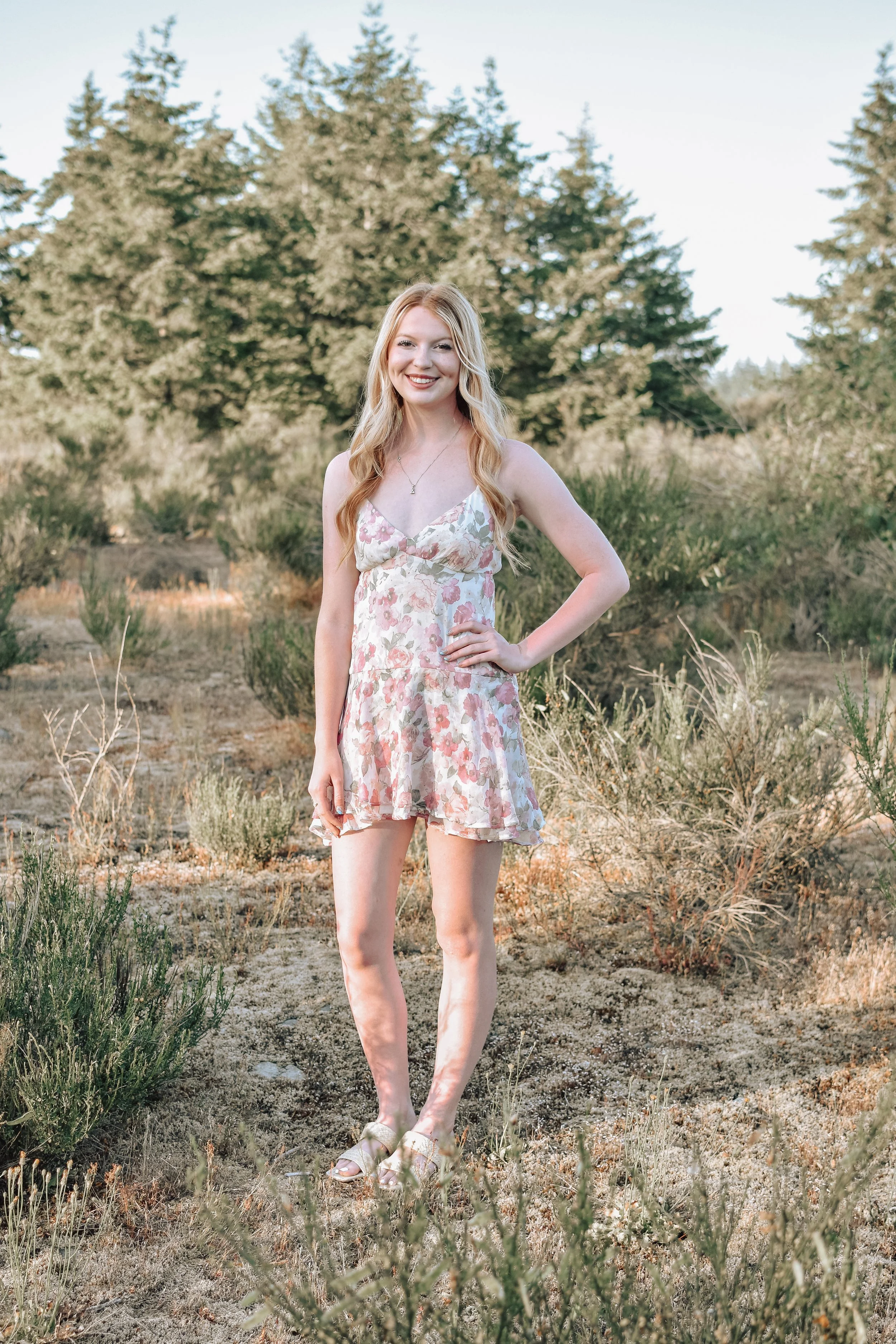 A young woman with long blonde hair in a floral dress standing outdoors in a dry, grassy area with trees in the background, smiling at the camera.