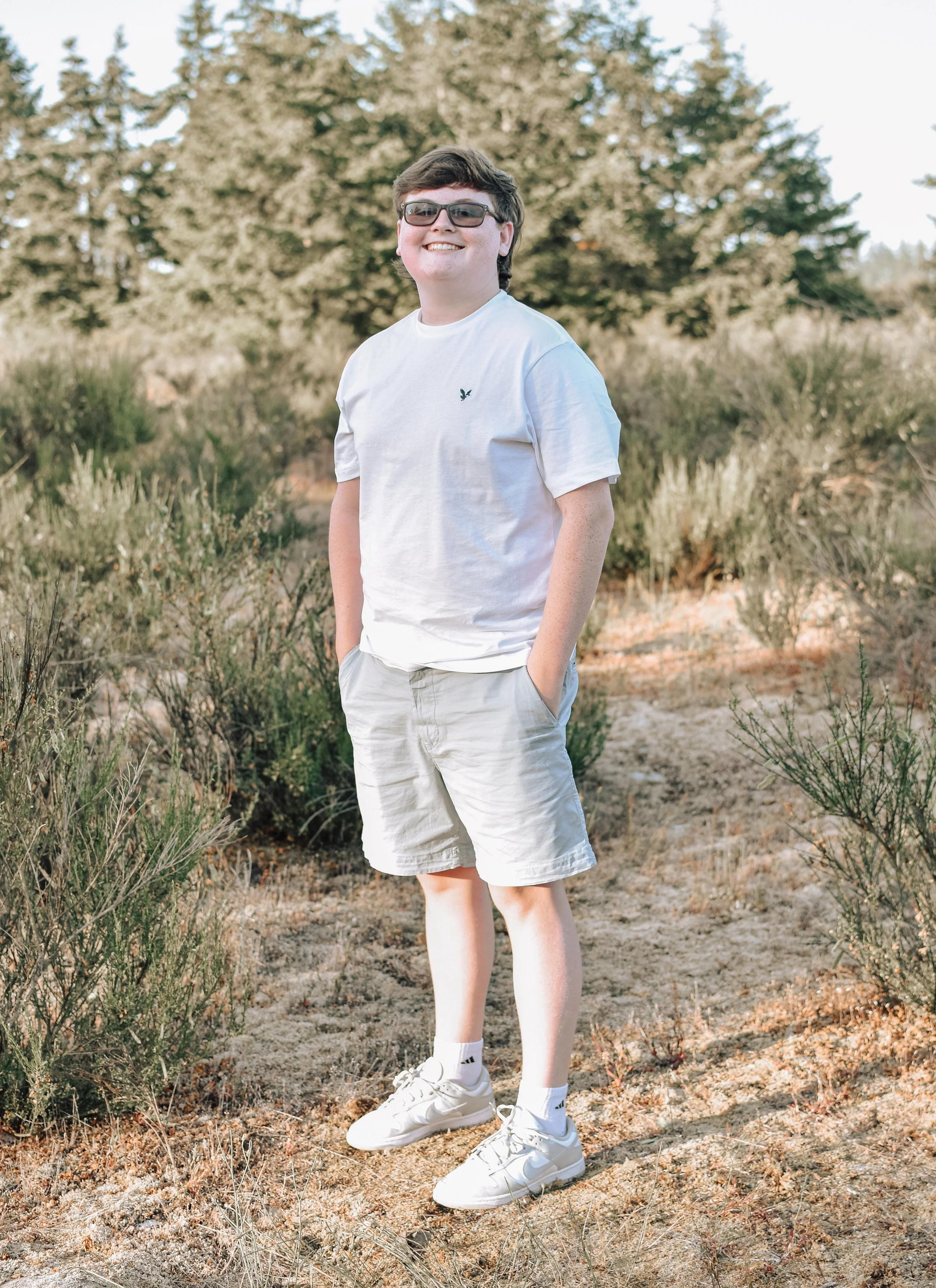 A smiling young man with glasses and dark hair in a white t-shirt, beige shorts, and white sneakers standing outdoors in a dry, shrub-covered landscape with some trees in the background.