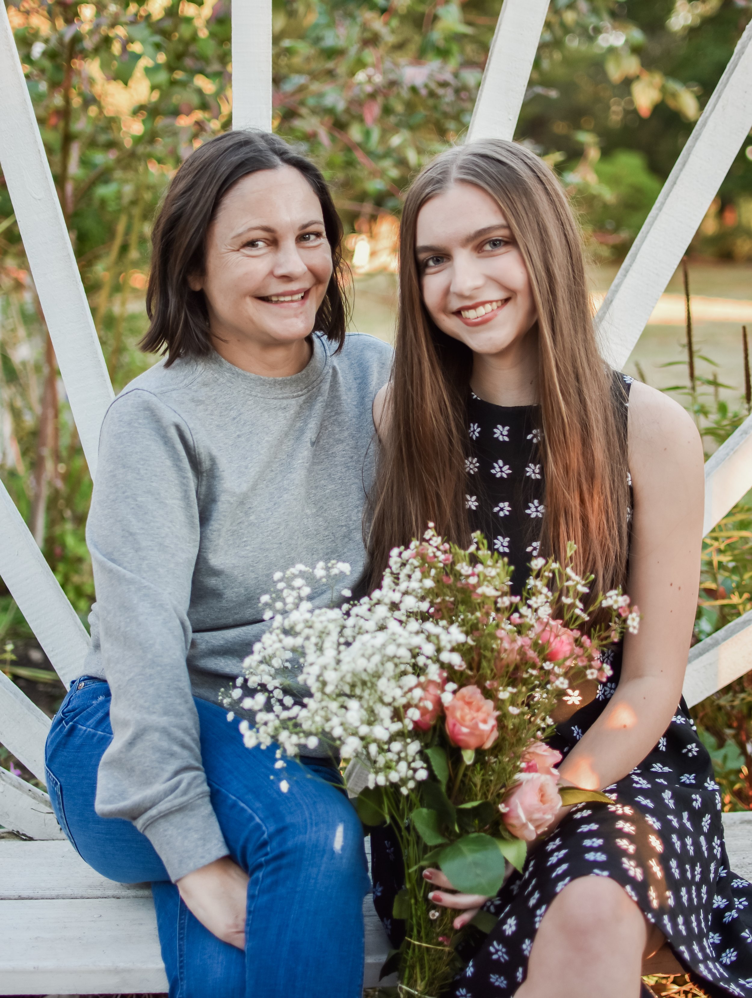 A woman with short dark hair and a young woman with long brown hair sitting on a white bench outdoors, holding a bouquet of pink and white flowers, surrounded by greenery and a white wooden structure.