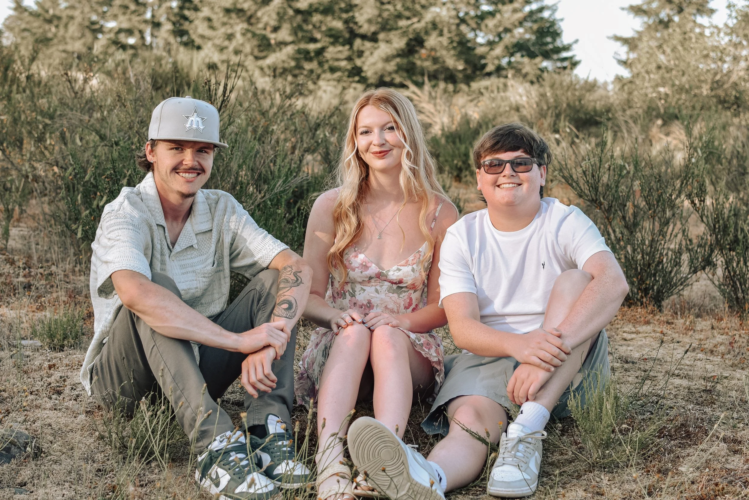 Three young people sitting outdoors on the ground, smiling, with bushes and trees in the background.