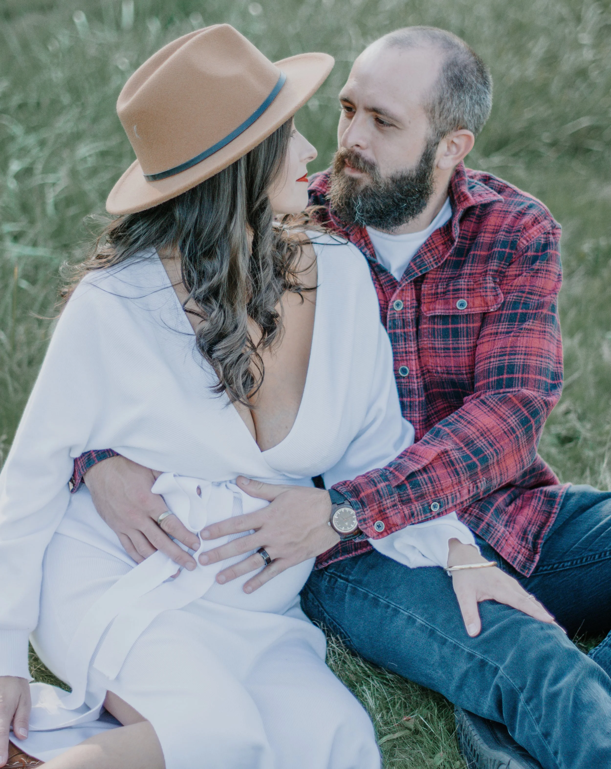 A man and woman sitting on grass, facing each other closely, with the man gently holding the woman's waist. The woman wears a wide-brimmed tan hat and a white dress, while the man sports a red plaid shirt, jeans, and a watch. They are outdoors in a n