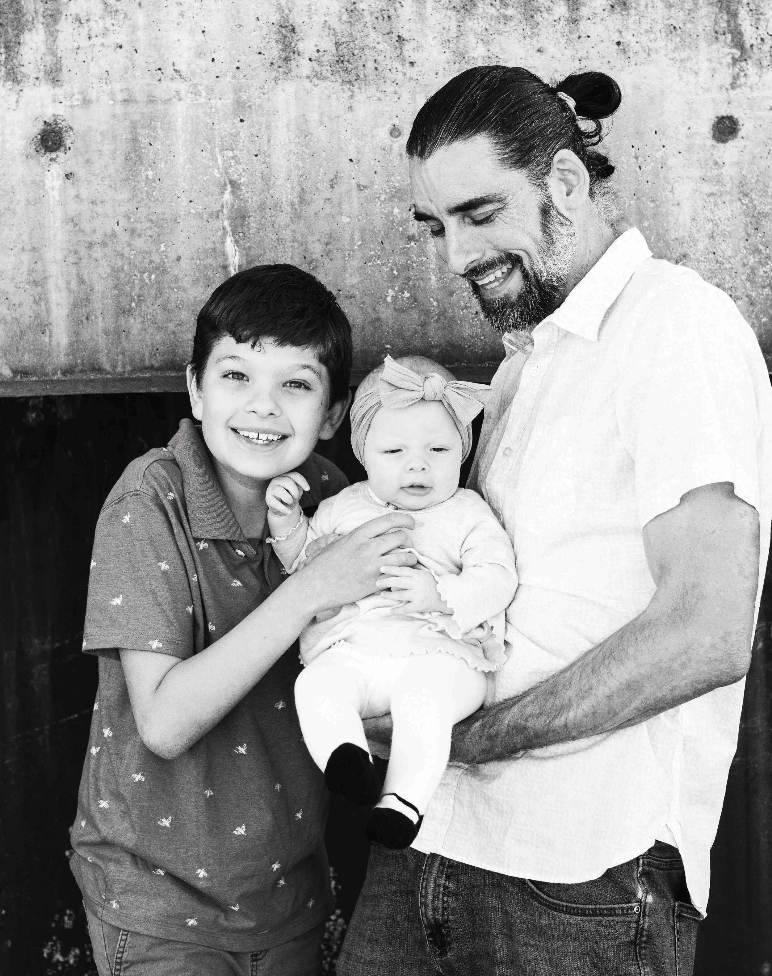 A family of three with a father, son, and baby girl smiling and looking at the camera against a textured wall.