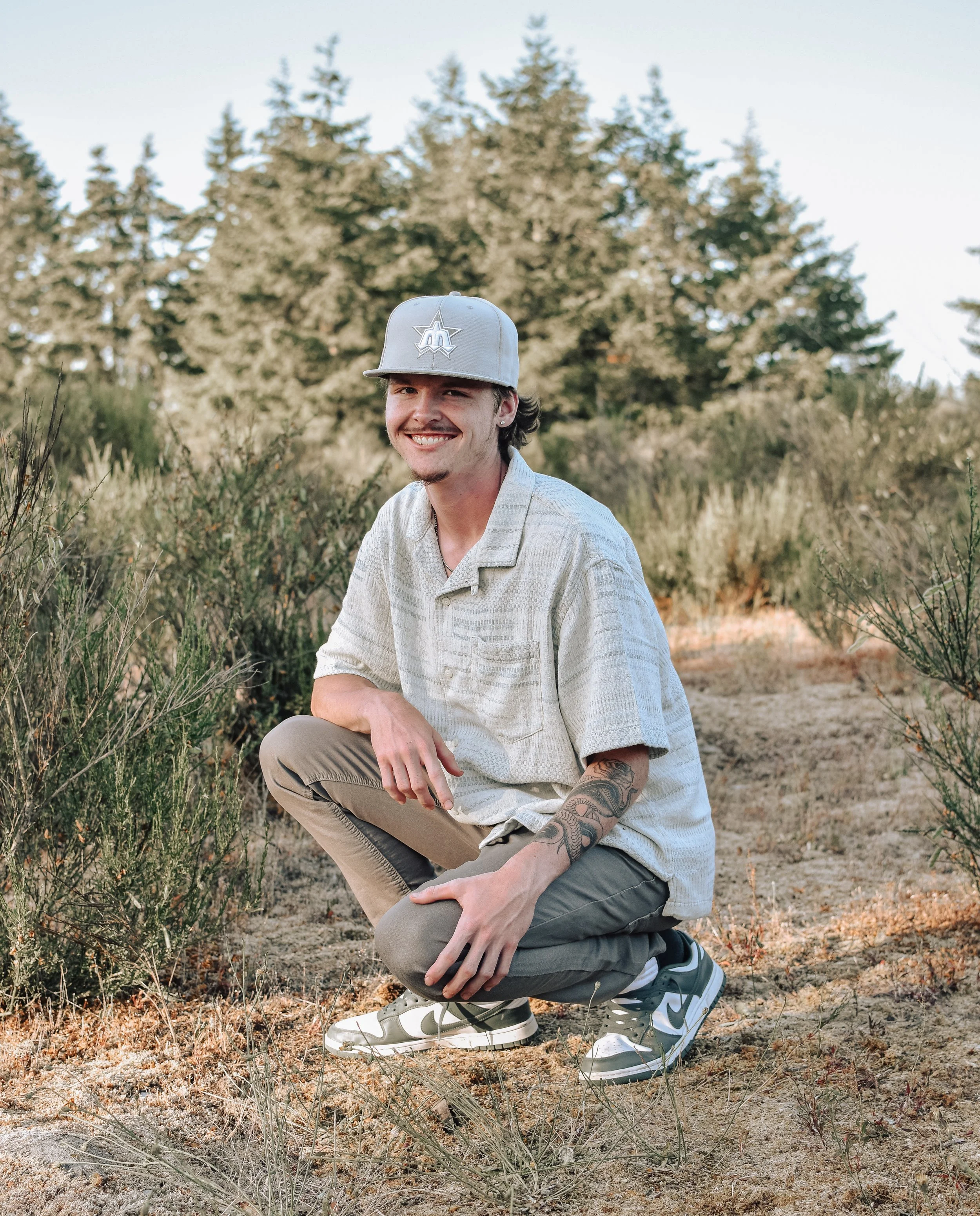 A young man with dark hair, facial hair, and tattoos on his right arm, squatting in a natural outdoor setting with bushes and trees in the background. He is smiling, wearing a light gray cap, a beige short-sleeve shirt, khaki pants, and Nike sneakers