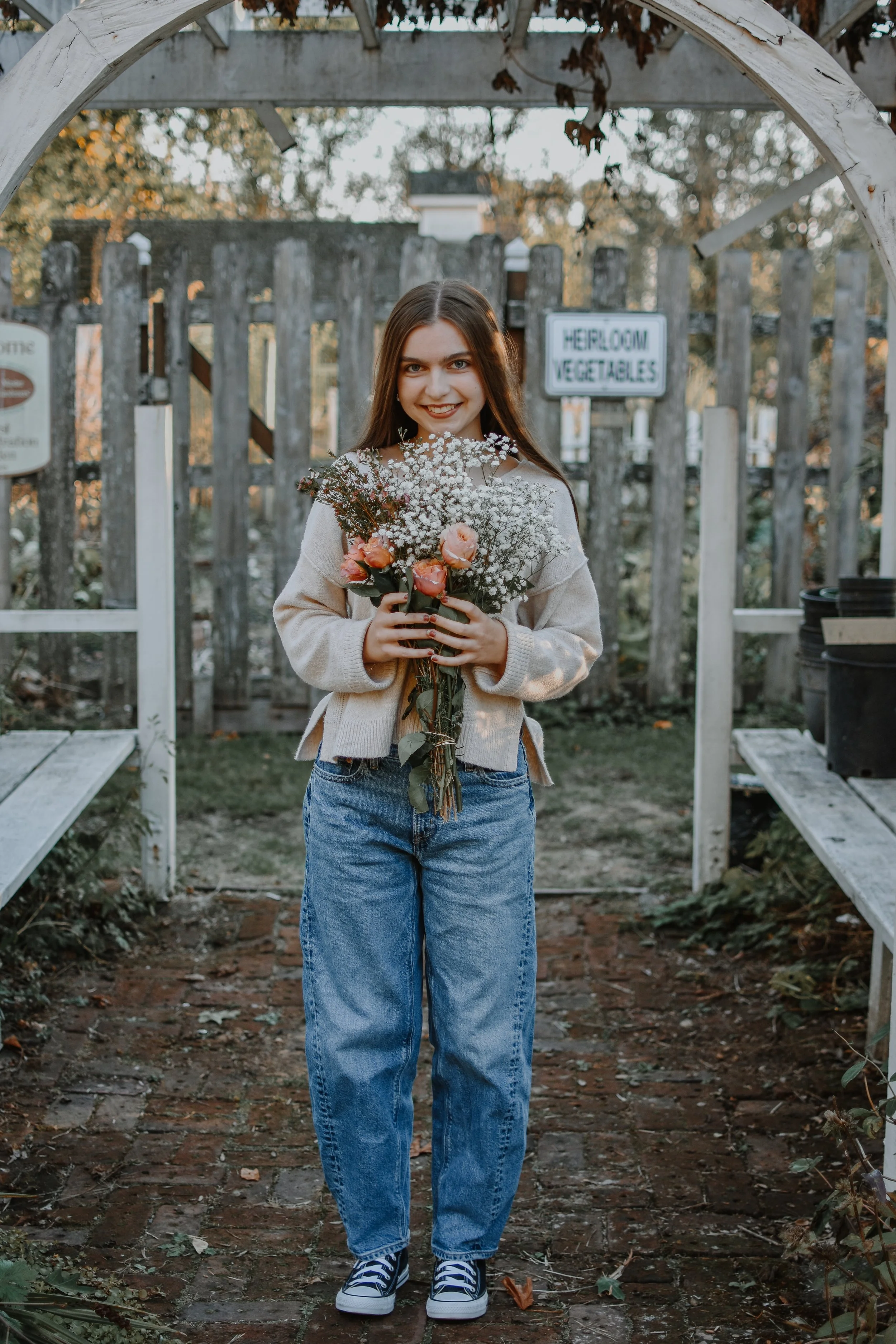 A young woman with long brown hair holding a bouquet of pink and white flowers, standing in a garden with a wooden fence and signs in the background.
