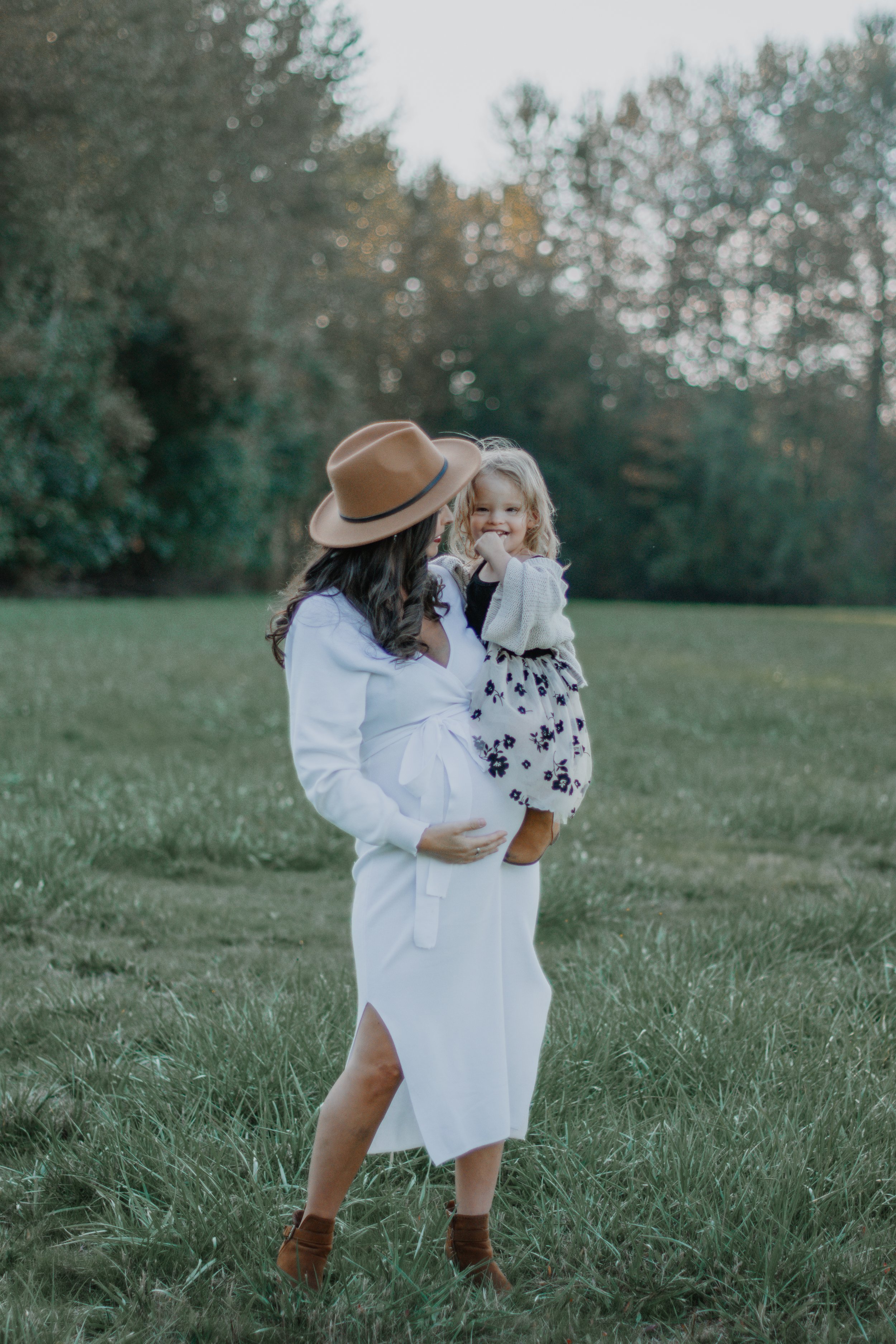 A woman holding a young girl outdoors in a grassy field with trees in the background. The woman wears a white dress, brown boots, and a wide-brimmed hat. The girl has blonde hair, a black top, and a floral skirt, smiling and touching her mouth.