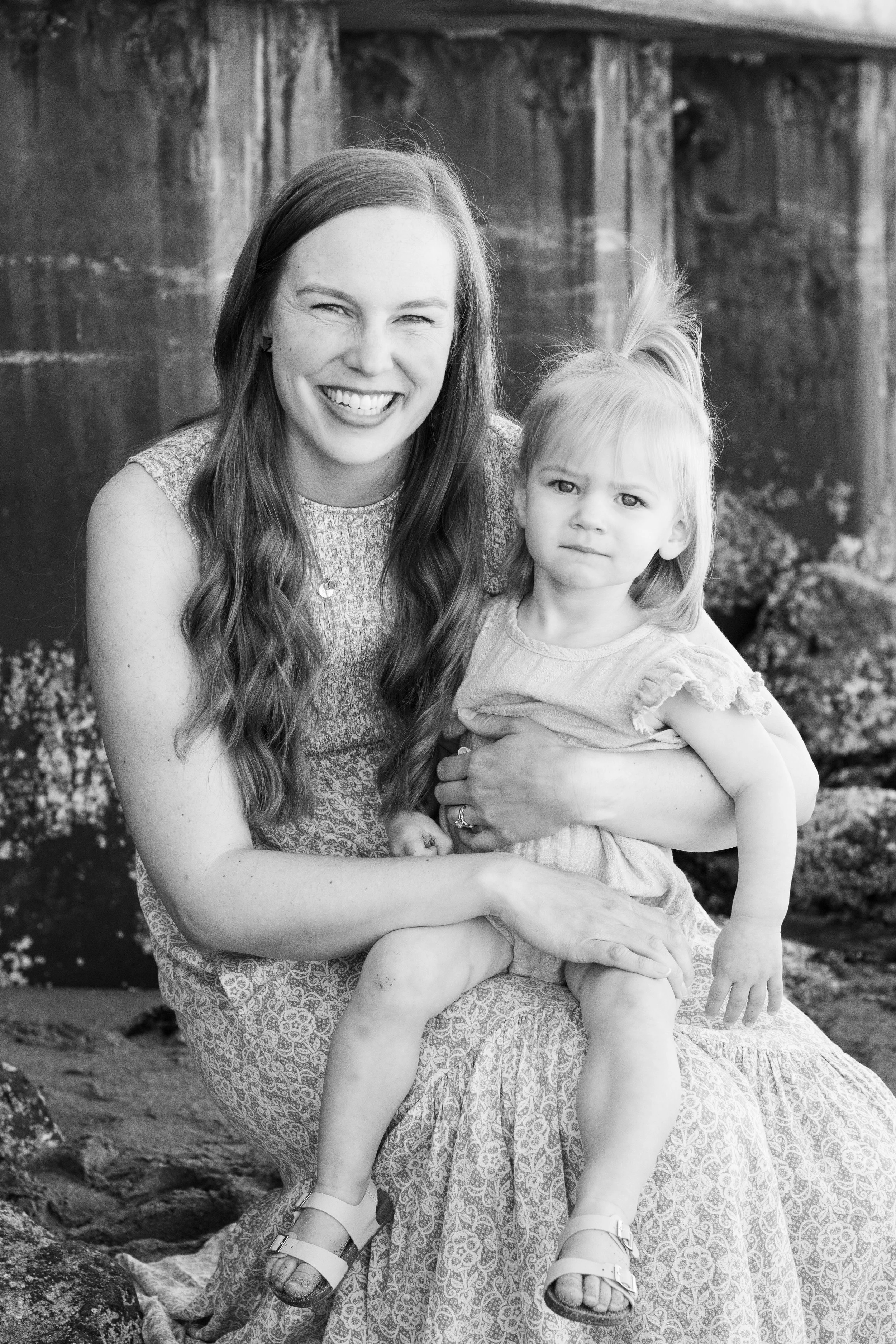 Black and white photo of a smiling woman with long wavy hair holding a serious young girl with long hair, outdoors near rocks and a wooden structure.
