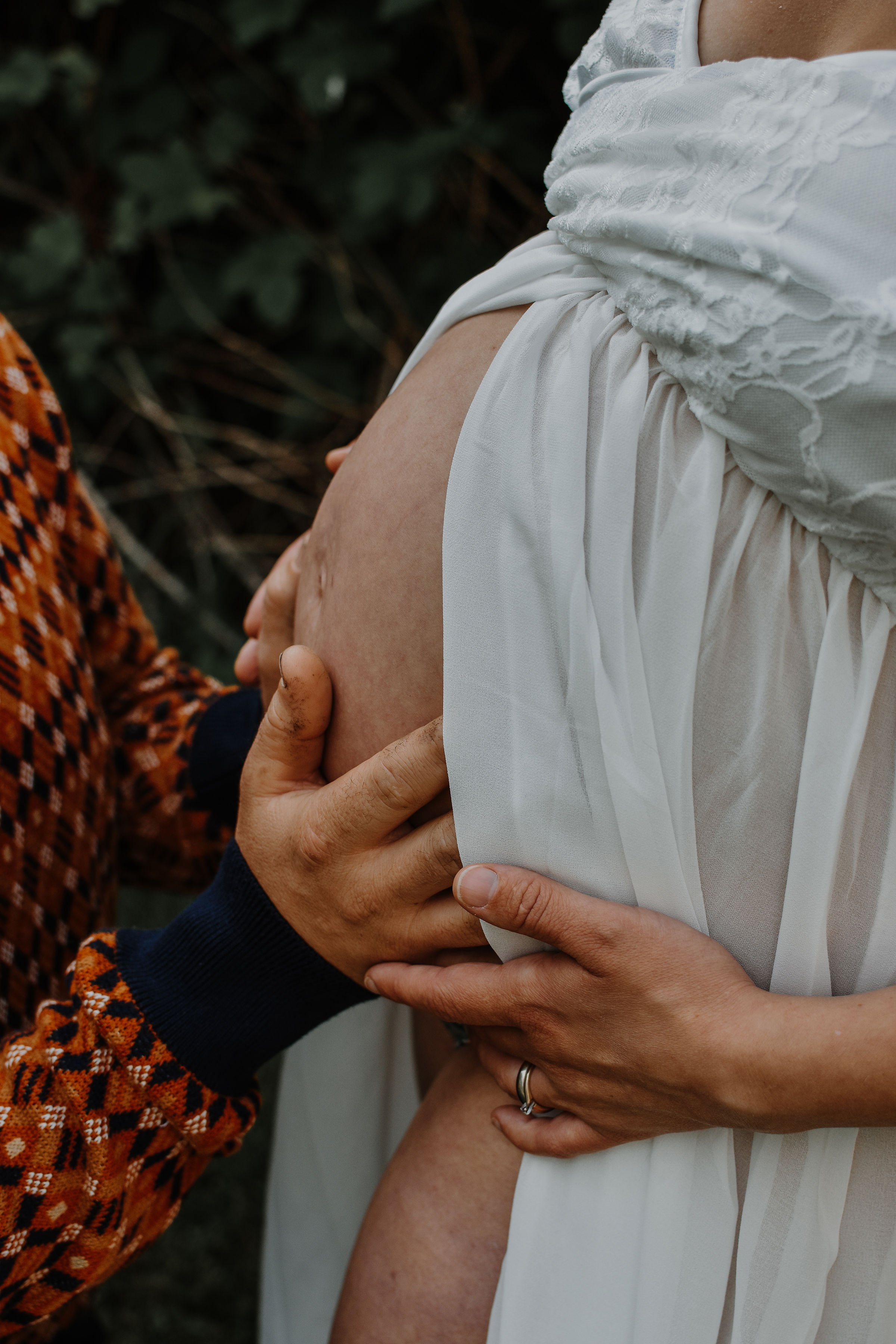 Person with a pregnant belly being gently held and touched by their partner's hands, outdoors.
