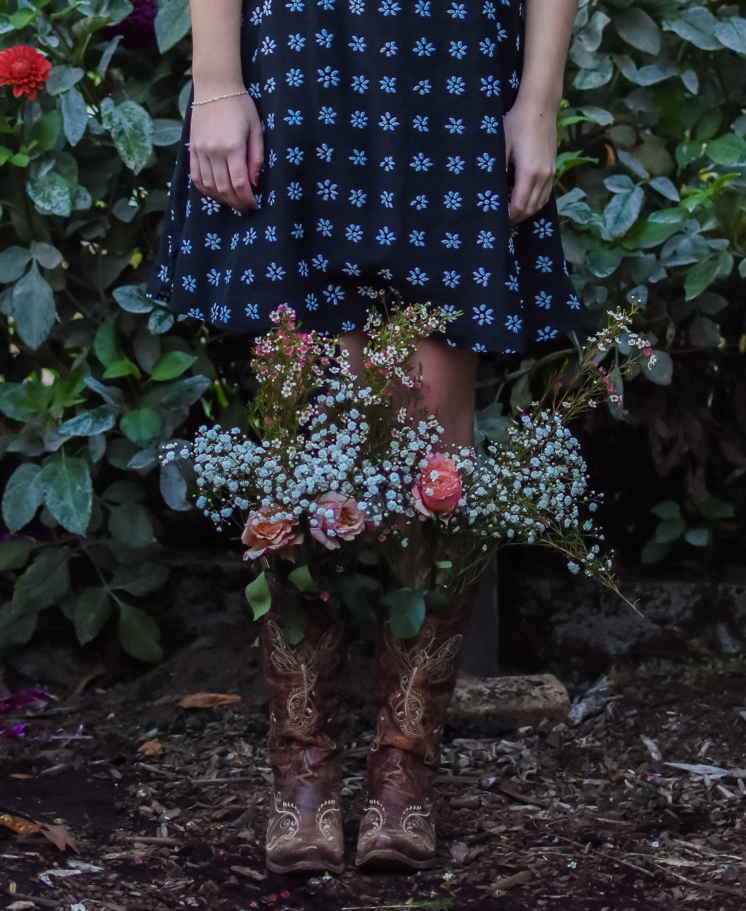 Person standing outdoors on dirt ground, wearing a navy blue dress with light blue flower pattern, cowboy boots with floral embroidery, holding a bouquet of mixed flowers including roses and baby's breath, with lush green foliage in the background.