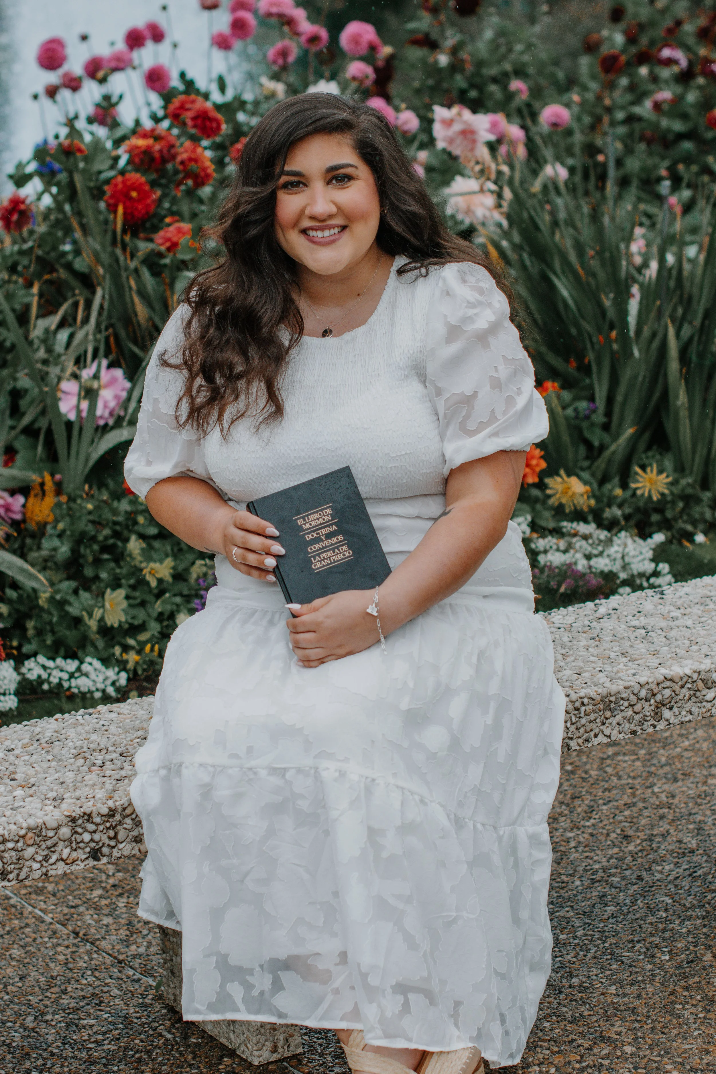 A young woman with long, curly dark hair, sitting on a stone bench outdoors, surrounded by colorful flowers. She is smiling, wearing a white dress with puffed sleeves, and holding a black book.