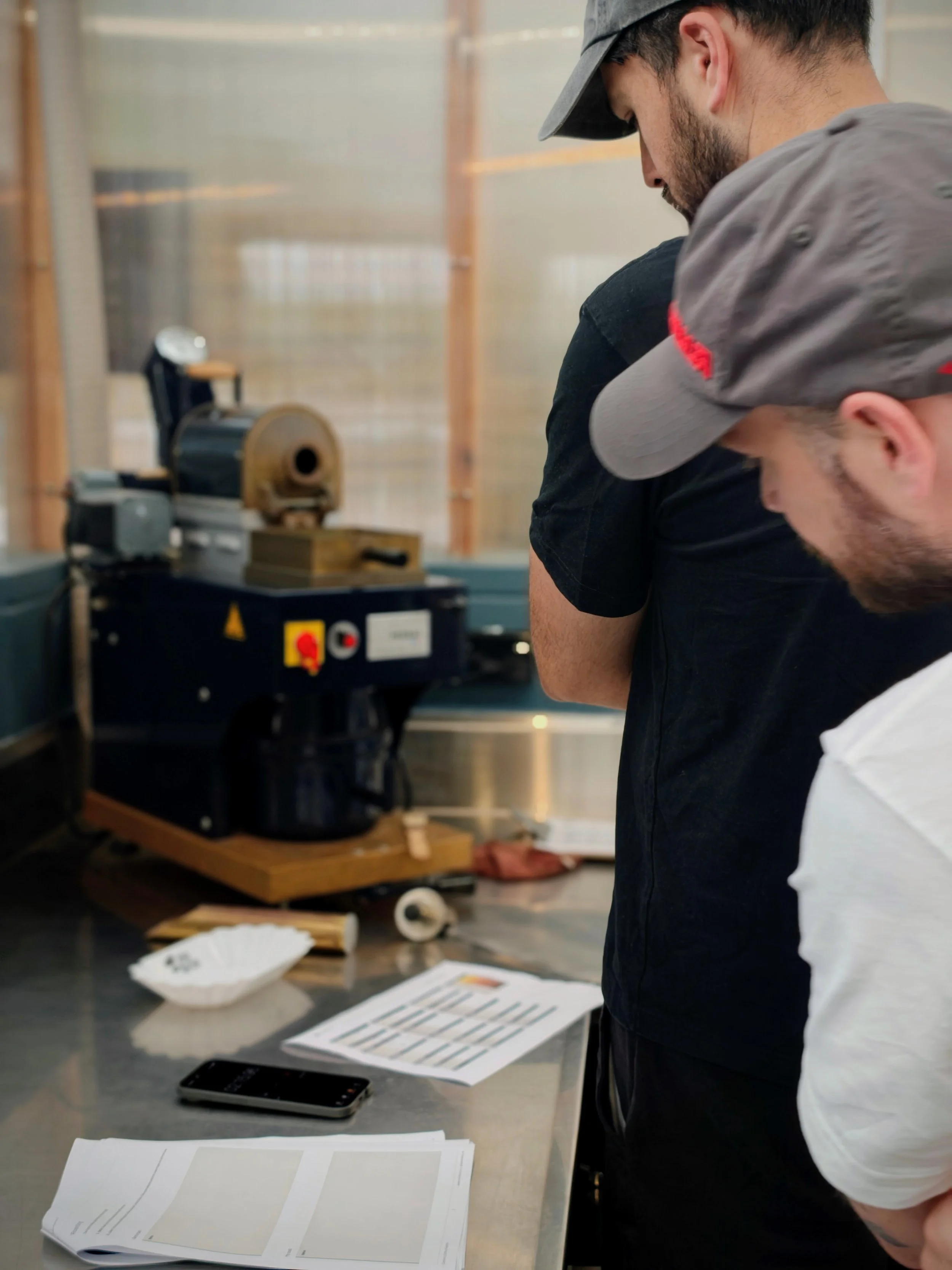 Two people reviewing a roast profile sheet at Common Coffee Roasters, with the Probat sample roaster visible in the background during a Sample Roasting 101 coffee class.
