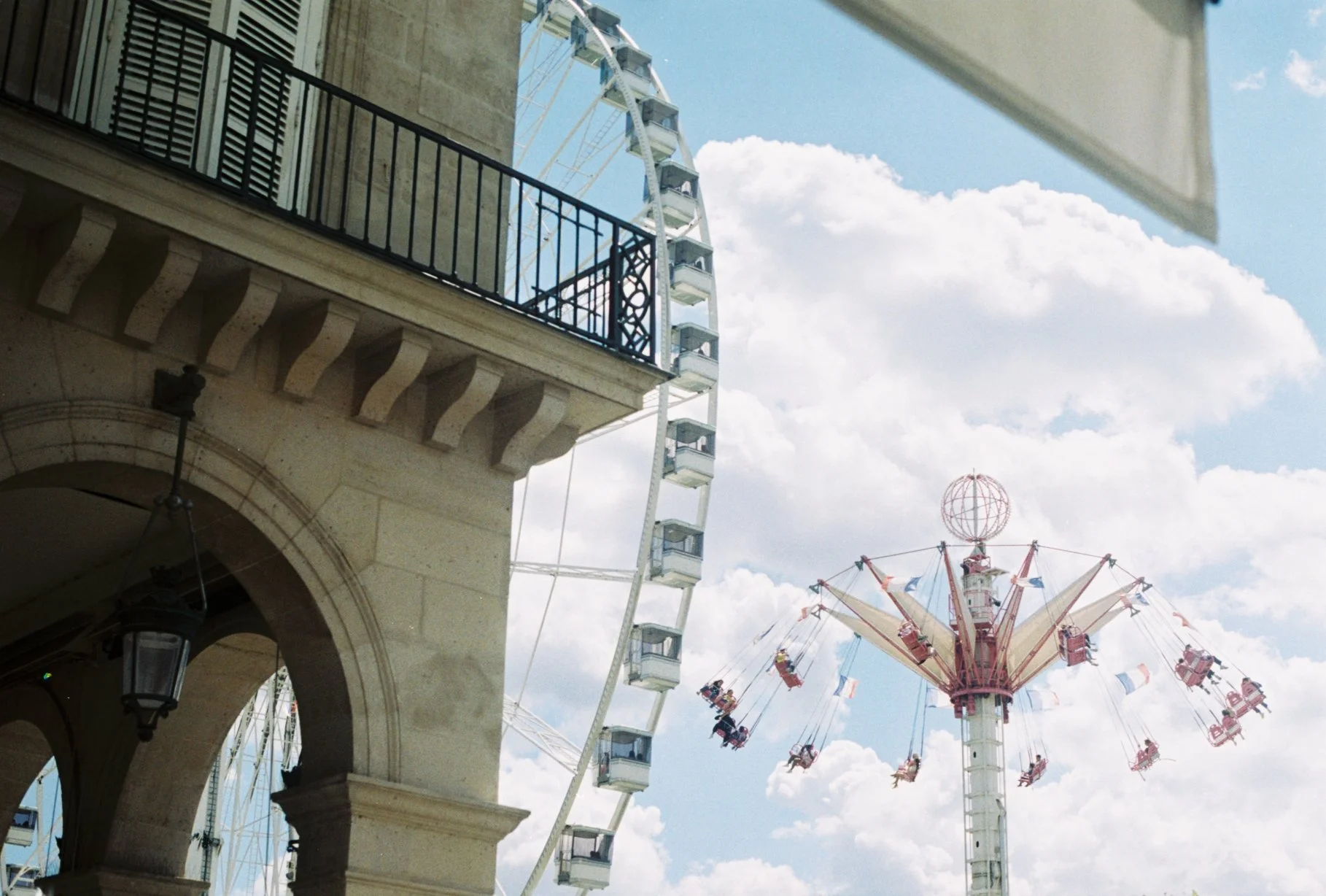 A Ferris wheel and a swing ride at an amusement park, with part of a building and balcony in the foreground, and cloudy sky in the background.